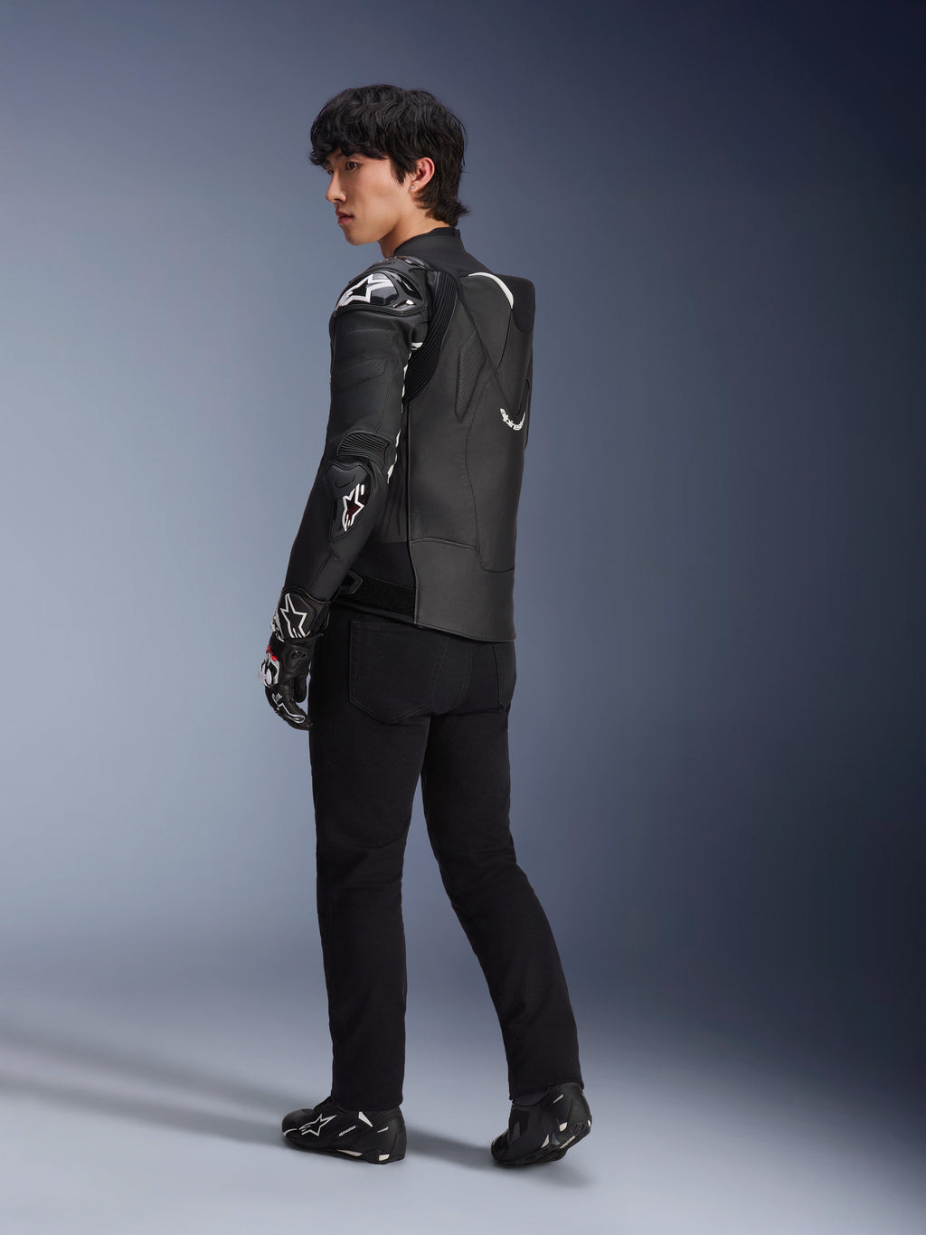 Male model wearing the Alpinestars Atem V5 Leather Jacket in Black and White, three-quarter rear view showing the aerodynamic speed hump and DFS RR protectors, styled with black pants, racing gloves, and boots, professional motorcycle racing apparel
