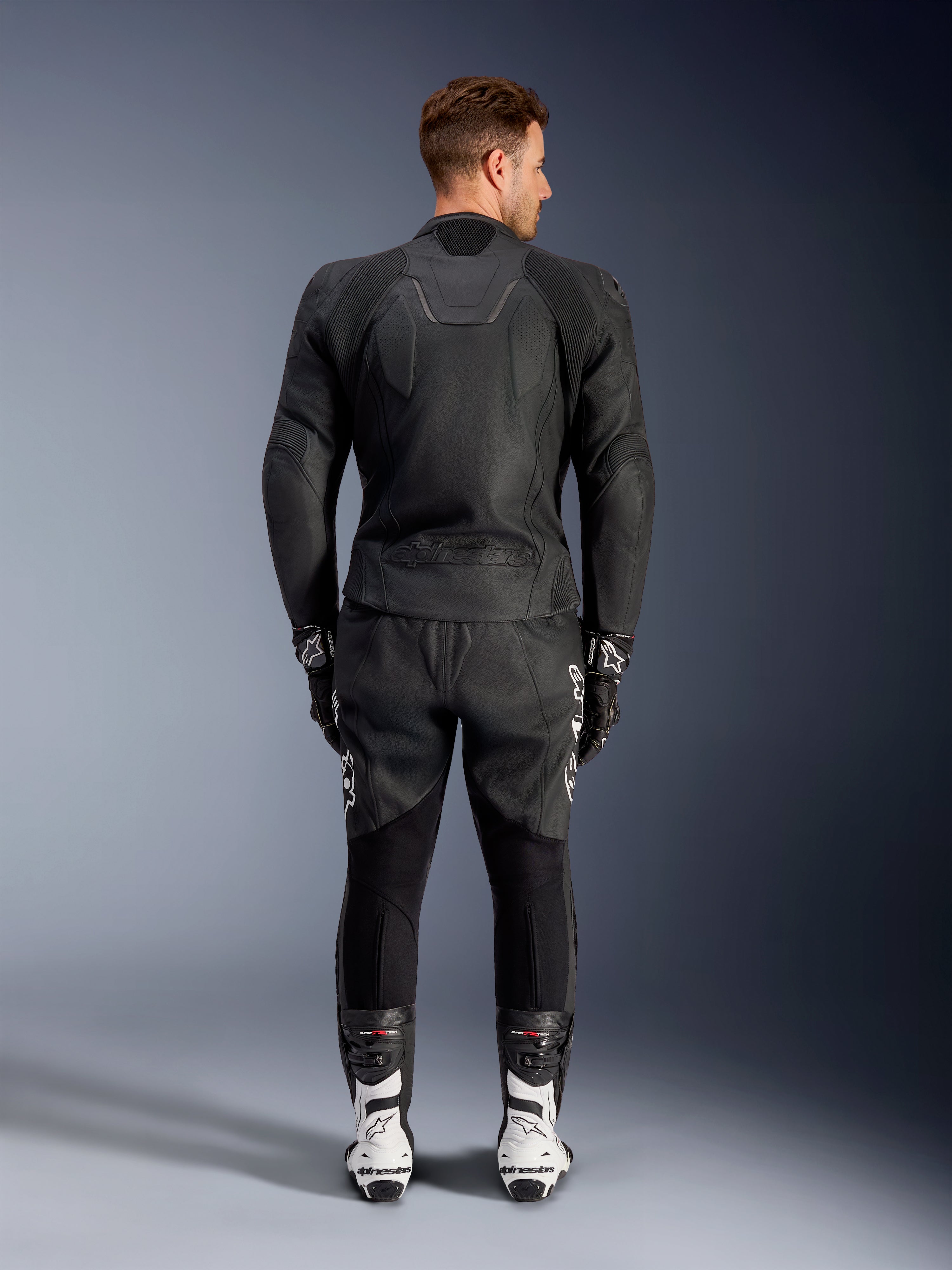 Vista traseira de um homem a usar o Casaco de Couro Caliber da Alpinestars em preto, com logótipo em relevo e painéis de couro perfurado, combinado com calças de motociclismo em couro preto, luvas pretas e botas de corrida brancas.
