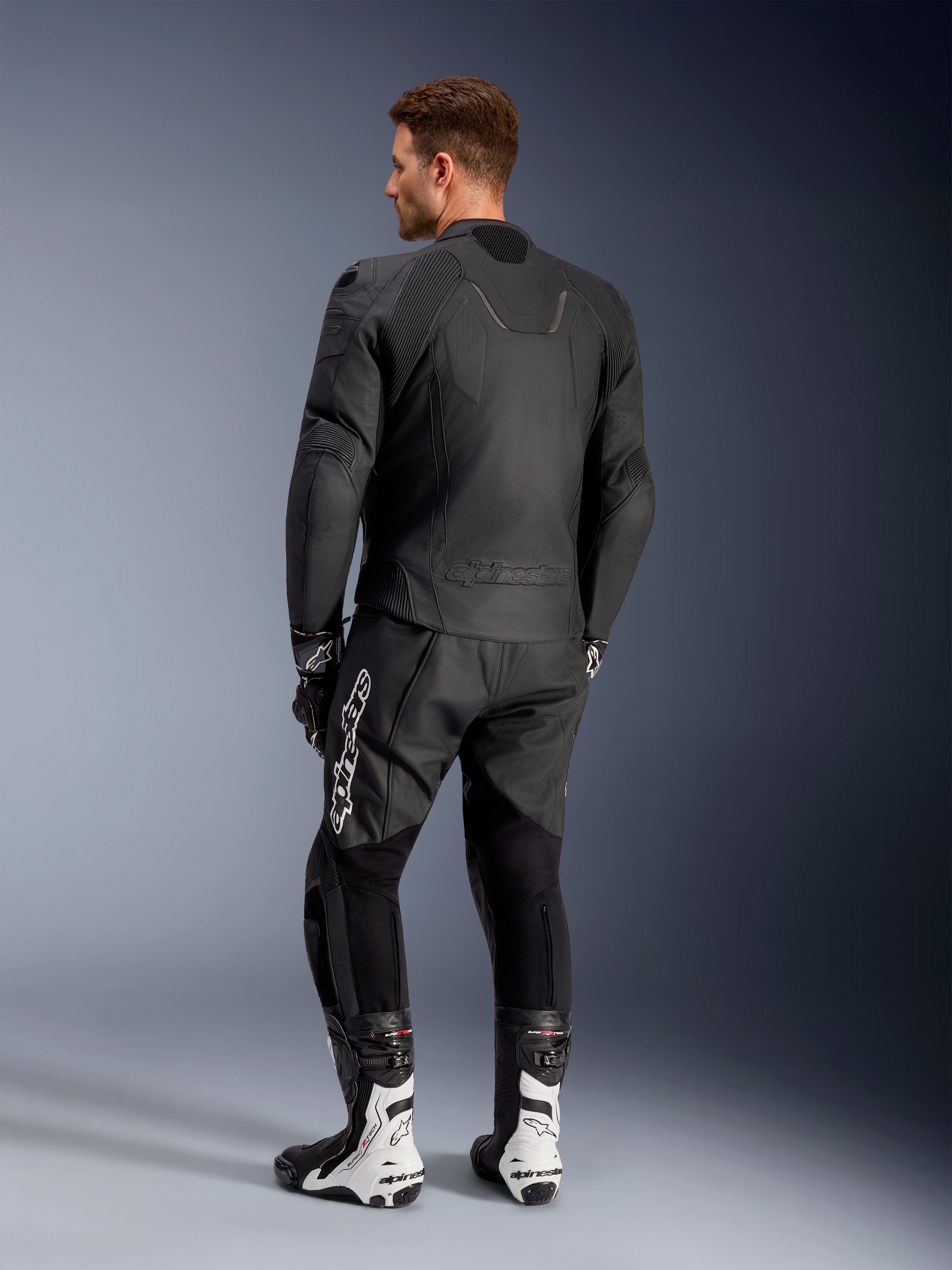 Casaco de Couro Alpinestars Caliber, casaco de motociclismo de estrada preto, vista traseira, com couro perfurado e inserções elásticas em acordeão na cintura e nos ombros, logótipo em relevo, usado com calças de couro pretas e botas de competição brancas.