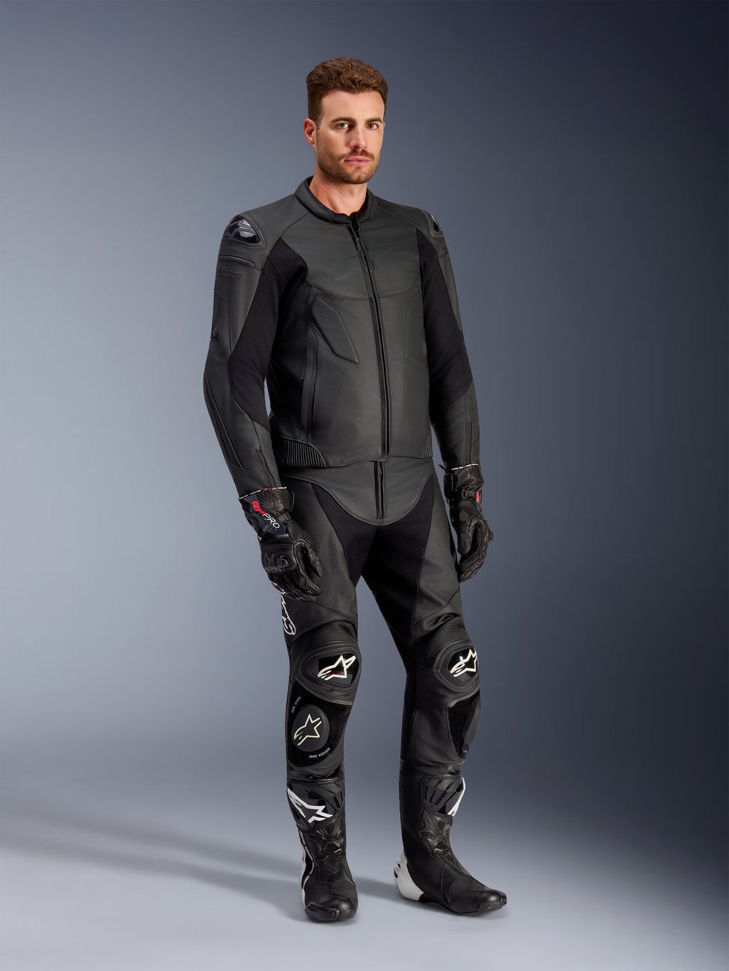 Modelo masculino a usar o casaco de couro Alpinestars Caliber em preto, combinado com calças de motociclismo de couro pretas a condizer, luvas de corrida e botas, apresentando um conjunto completo de equipamento desportivo de estrada.