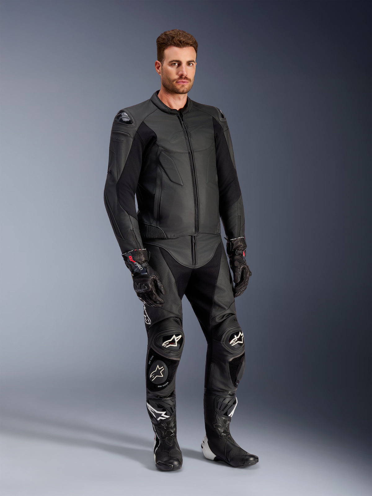 Modelo masculino a usar o casaco de couro Alpinestars Caliber em preto, combinado com calças de motociclismo de couro pretas a condizer, luvas de corrida e botas, apresentando um conjunto completo de equipamento desportivo de estrada.