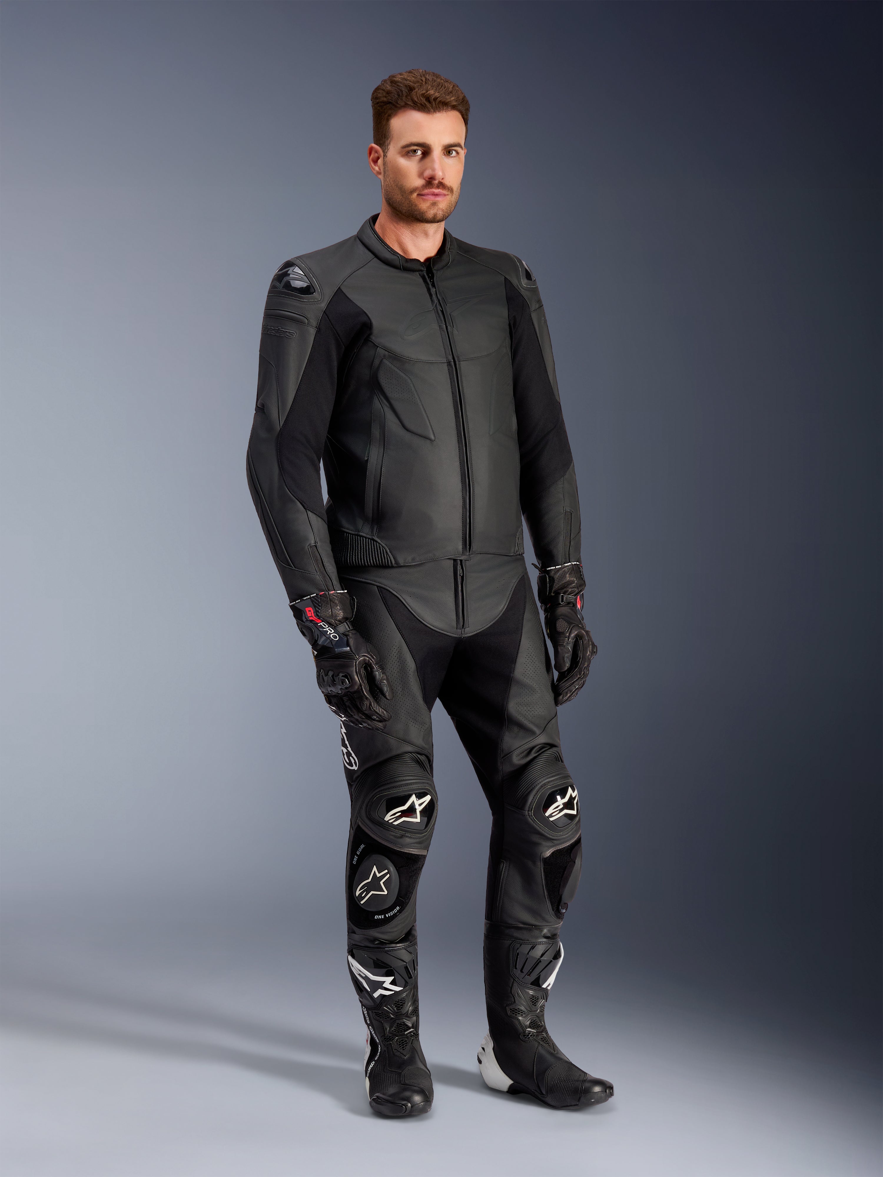 Modelo masculino a usar o casaco de couro Alpinestars Caliber em preto, combinado com calças de motociclismo de couro pretas a condizer, luvas de corrida e botas, apresentando um conjunto completo de equipamento desportivo de estrada.