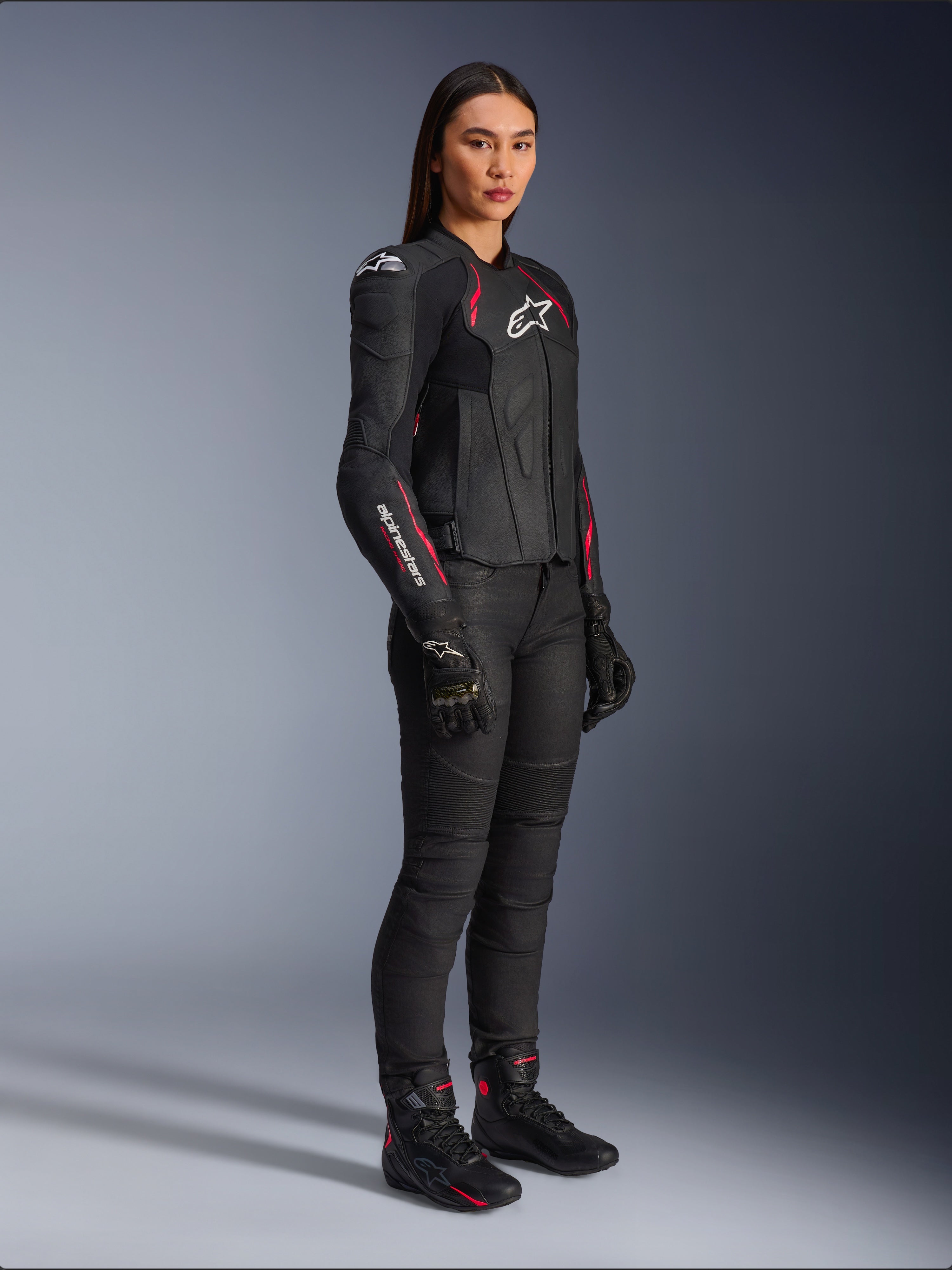 Casaco de Couro Stella Dusk Feminino da Alpinestars, Preto, Rosa Diva e Branco, usado por uma modelo com calças e botas de motociclismo pretas, apresentando uma construção em couro elegante com detalhes em rosa e logótipo branco.
