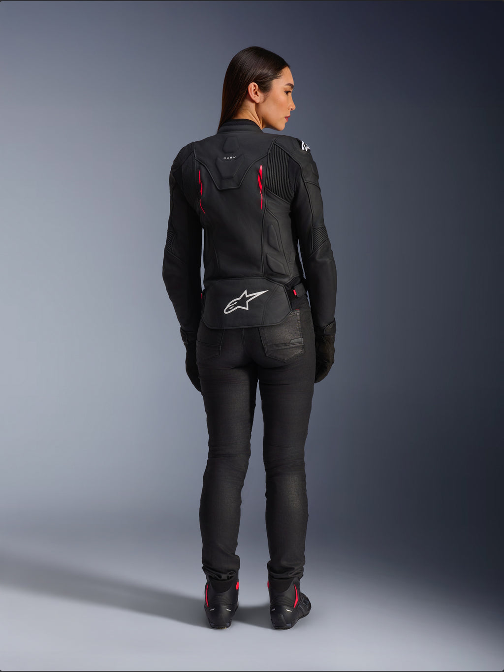 Casaco de Couro Alpinestars Stella Dusk, vista traseira, couro preto com detalhes em Diva Pink e branco, apresenta logótipo Alpinestars branco na parte inferior das costas e pormenores de ventilação cor-de-rosa, usado com calças e botas de condução pretas, equipamento de motociclismo desportivo premium para mulher