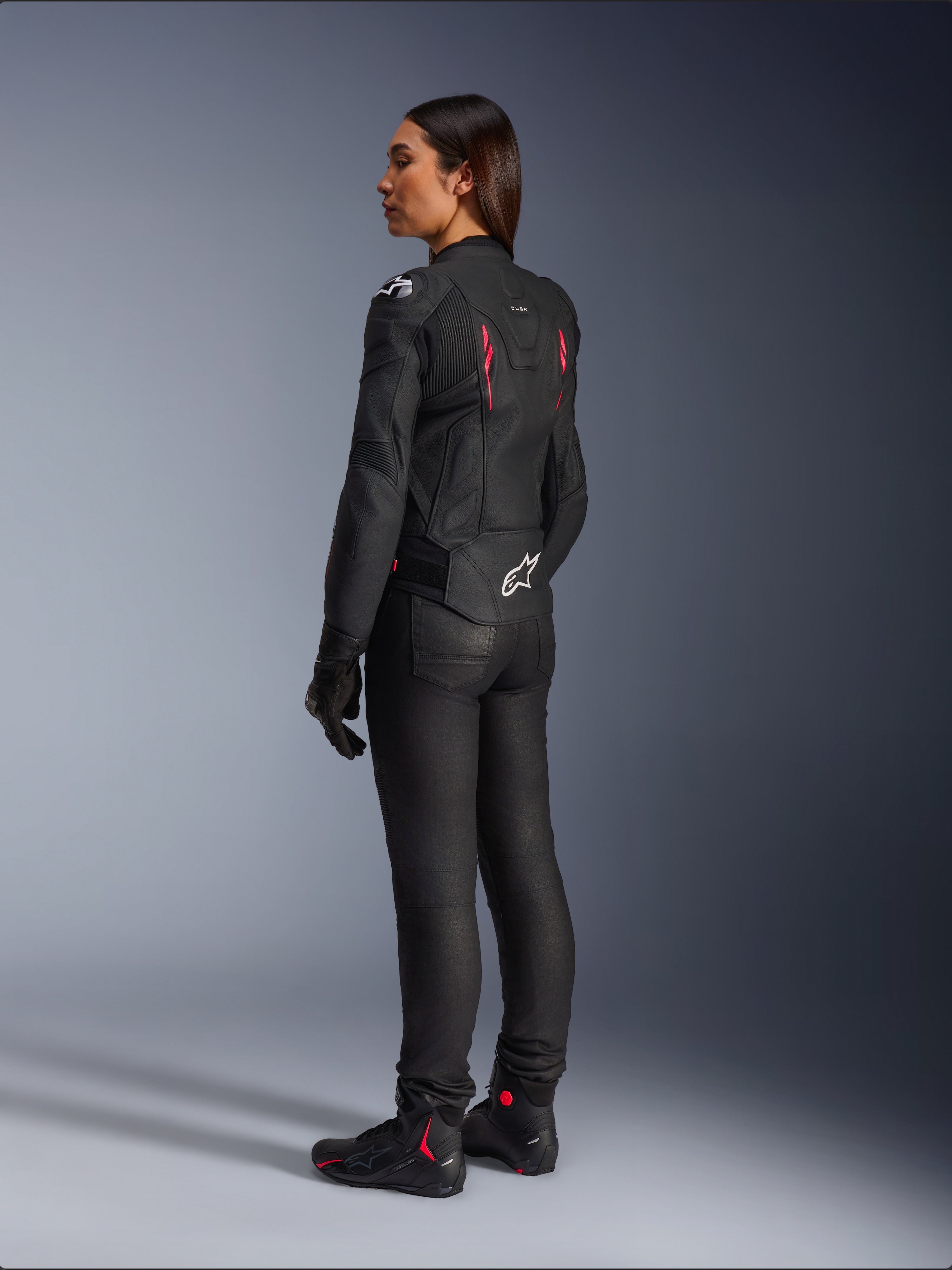 Casaco de Couro Stella Dusk para Mulher, Black Diva Pink White, vista traseira de três quartos numa motociclista, usado com calças de condução pretas e botas pretas, com construção em couro elegante, detalhes em rosa e logótipo Alpinestars branco na parte inferior das costas.