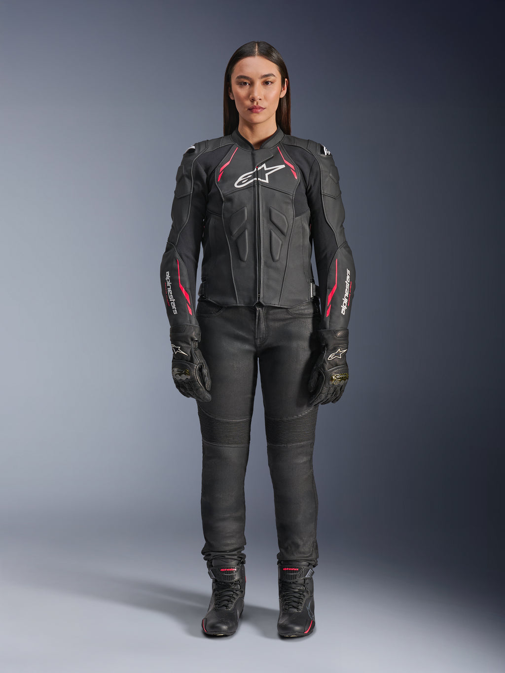 Uma mulher a usar o casaco de couro Alpinestars Stella Dusk em Black Diva Pink White, com um logótipo branco no peito e detalhes cor-de-rosa nos ombros e mangas, combinado com calças, luvas e botas de motociclismo pretas.