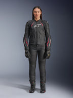 Uma mulher a usar o casaco de couro Alpinestars Stella Dusk em Black Diva Pink White, com um logótipo branco no peito e detalhes cor-de-rosa nos ombros e mangas, combinado com calças, luvas e botas de motociclismo pretas.