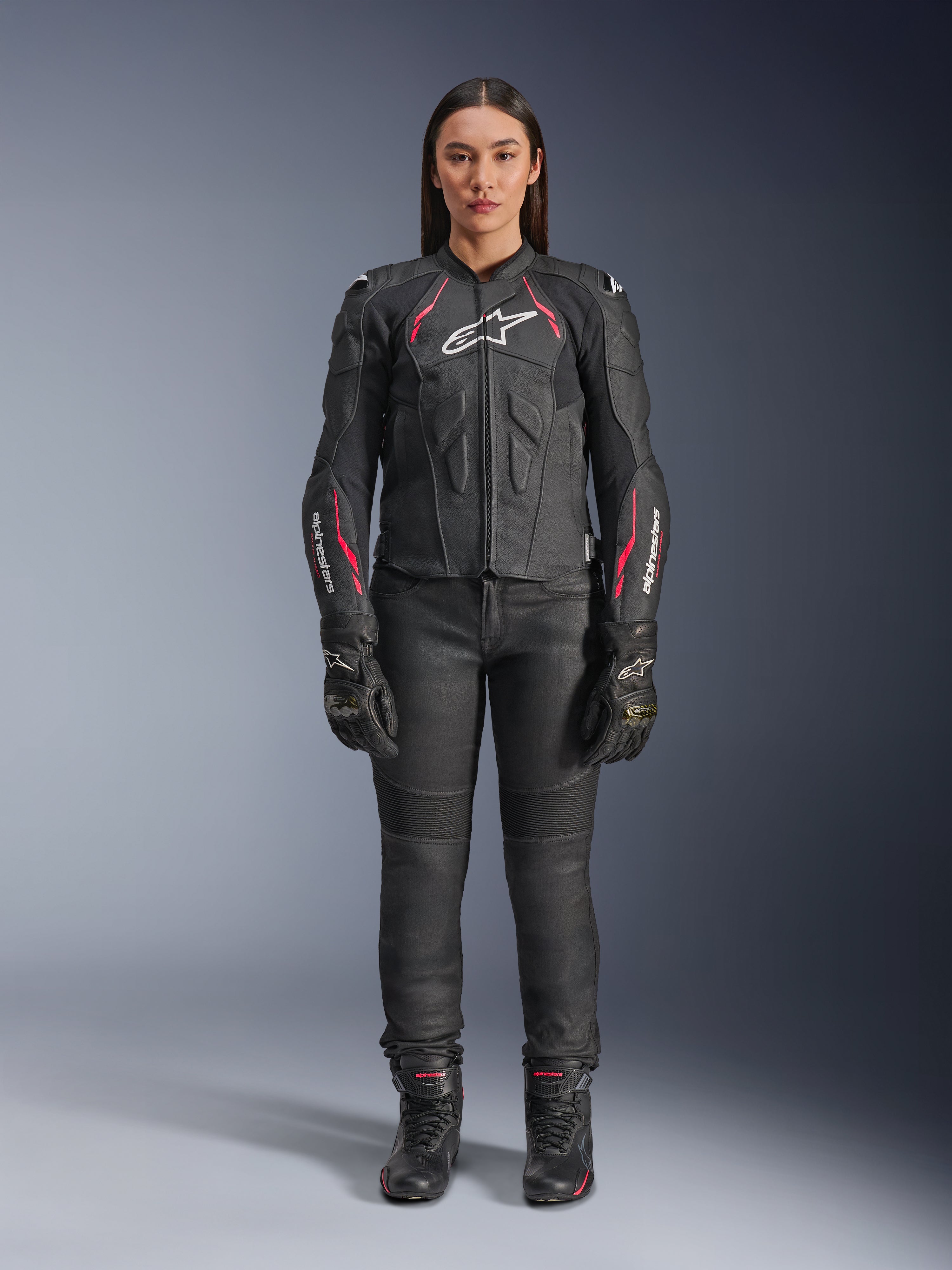 Uma mulher a usar o casaco de couro Alpinestars Stella Dusk em Black Diva Pink White, com um logótipo branco no peito e detalhes cor-de-rosa nos ombros e mangas, combinado com calças, luvas e botas de motociclismo pretas.