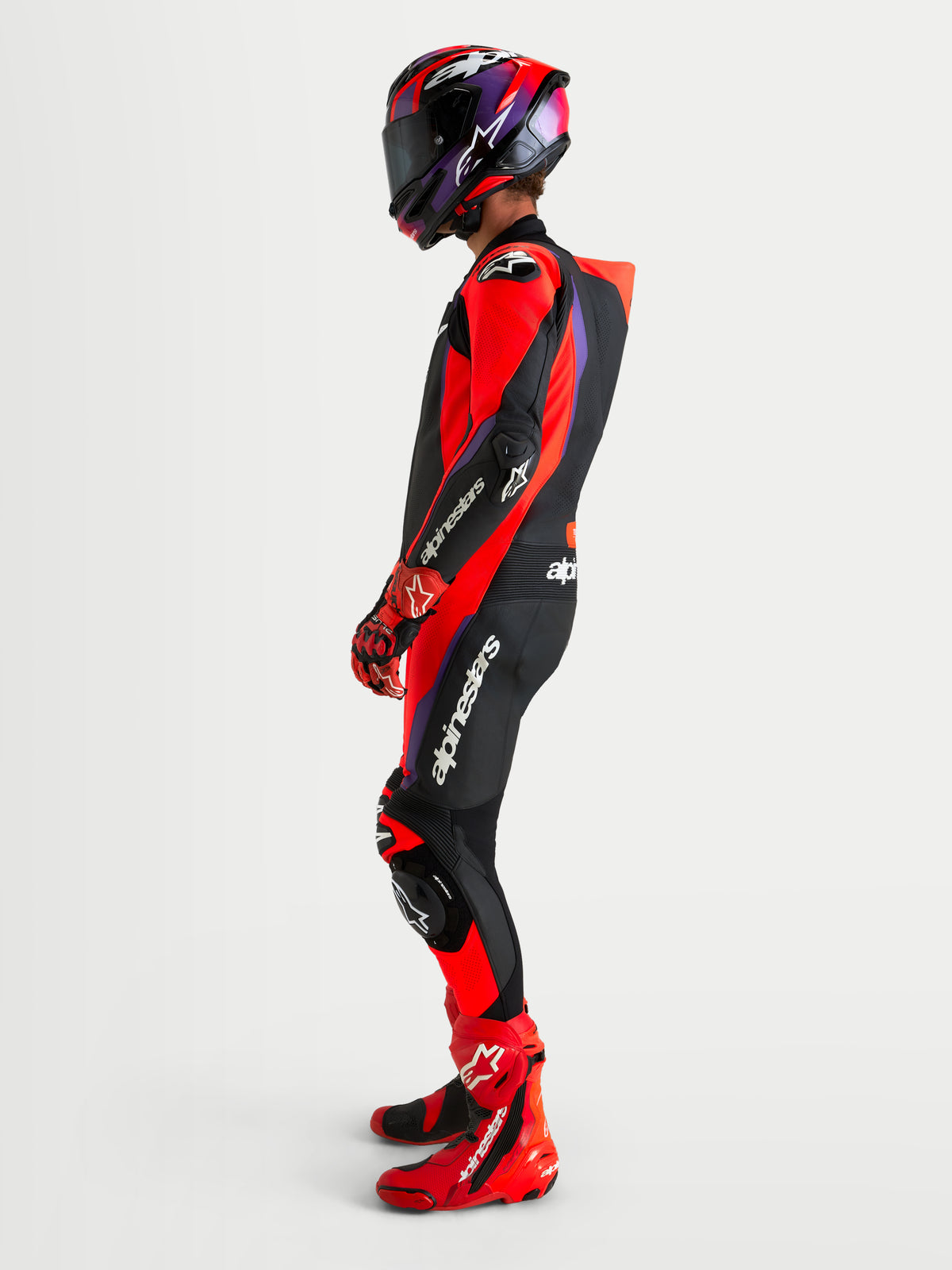 Alpinestars Missile V3 macacão de couro para corrida de moto para homem, preto, vermelho e roxo, com corcova aerodinâmica e proteção DFS ST, vista lateral num modelo com capacete, luvas e botas