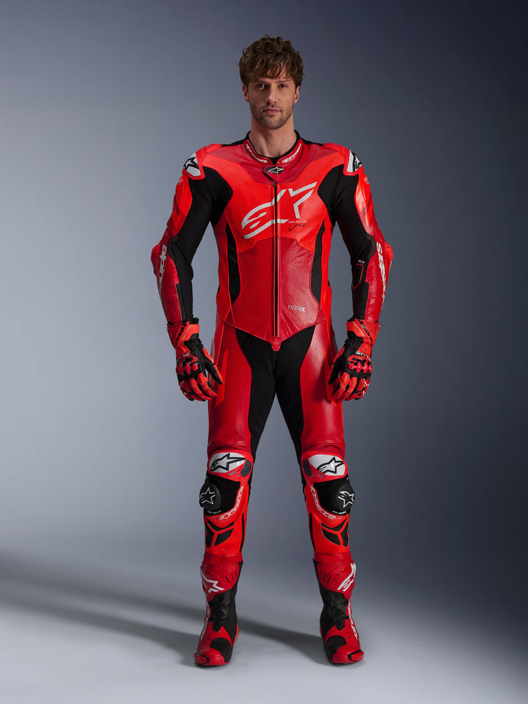 Modelo a usar o Fato de Pele Alpinestars GP Plus V4 Sprint 1PC, em Vermelho Fluo, Vermelho Médio e Branco, com detalhes em preto e branco, acompanhado por luvas e botas de corrida a condizer, design desportivo com logótipo grande no peito e painéis de pele perfurada para ventilação.
