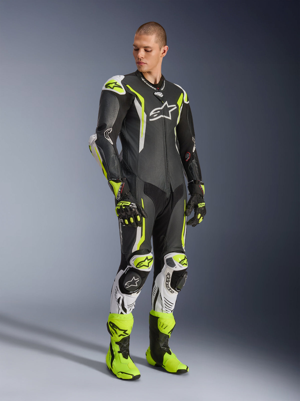 Fato de Couro Alpinestars GP Tech V5 de uma peça, preto, branco e amarelo fluo, vestido por um modelo com luvas e botas de corrida a condizer, equipamento profissional de corrida de motociclismo com desempenho aerodinâmico e proteções