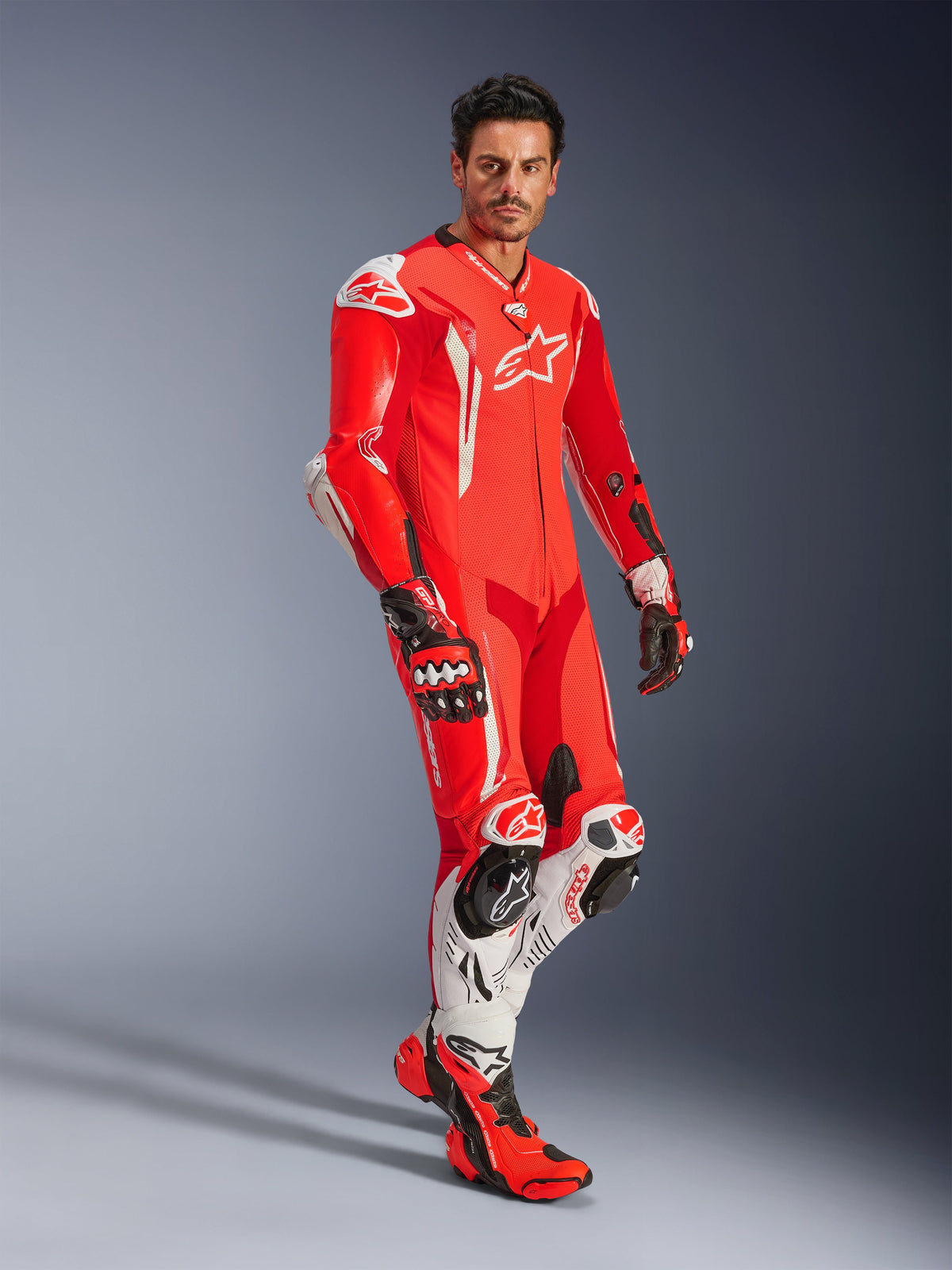 Homem com o Fato de Couro Alpinestars GP Tech V5 de uma peça, em Vermelho Fluo, Vermelho Vivo e Branco, combinado com luvas e botas de competição, equipamento profissional de motociclismo com corcova aerodinâmica e proteção integrada.