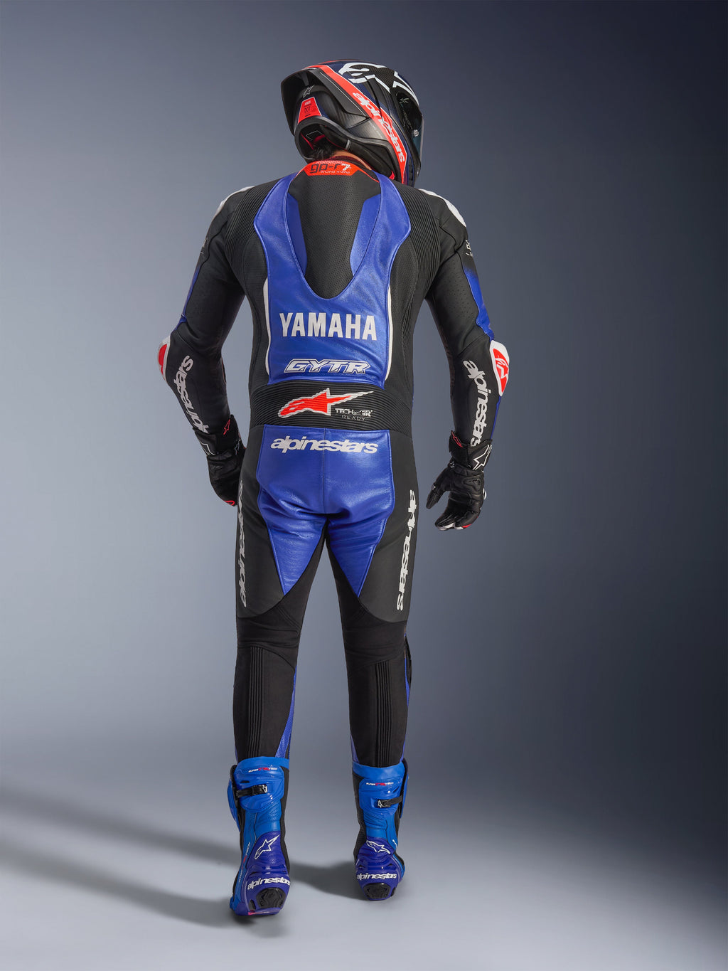 Fato de couro Alpinestars Yamaha GP-R7 1PC, vista traseira de um piloto de motociclismo com equipamento de corrida completo, Azul Metálico e Preto com pormenores em Branco e Vermelho, destacando os logótipos Yamaha e GYTR na corcunda aerodinâmica, botas azuis e capacete de corrida.