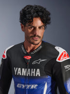 Retrato em grande plano de um homem a usar o fato de couro Alpinestars Yamaha GP-R7 1PC, azul metálico e preto com logótipos YAMAHA e GYTR brancos, couro perfurado, protetores de ombro brancos com logótipos de estrela vermelhos, Tech-Air Ready, equipamento profissional de motociclismo