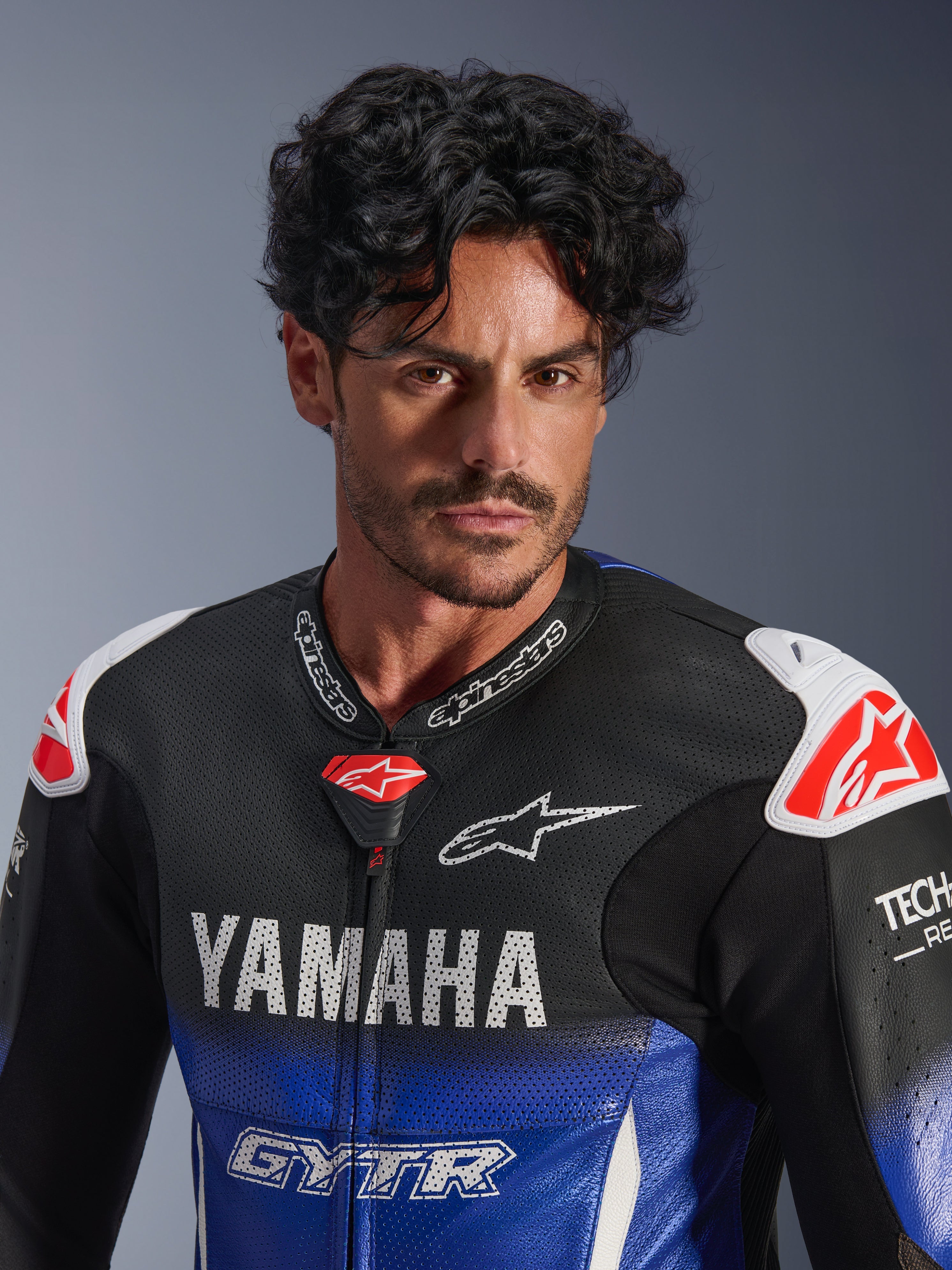 Retrato em grande plano de um homem a usar o fato de couro Alpinestars Yamaha GP-R7 1PC, azul metálico e preto com logótipos YAMAHA e GYTR brancos, couro perfurado, protetores de ombro brancos com logótipos de estrela vermelhos, Tech-Air Ready, equipamento profissional de motociclismo