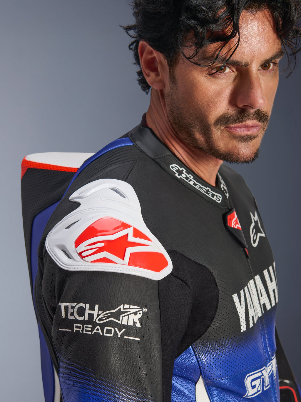Fato de Couro Yamaha GP-R7 1PC, Azul Metálico, Preto, Branco e Vermelho, vista aproximada do ombro e do peito, com proteção de ombro DFS branca com logótipo vermelho, corcunda aerodinâmica e marcação Tech-Air Ready na manga de couro perfurado.