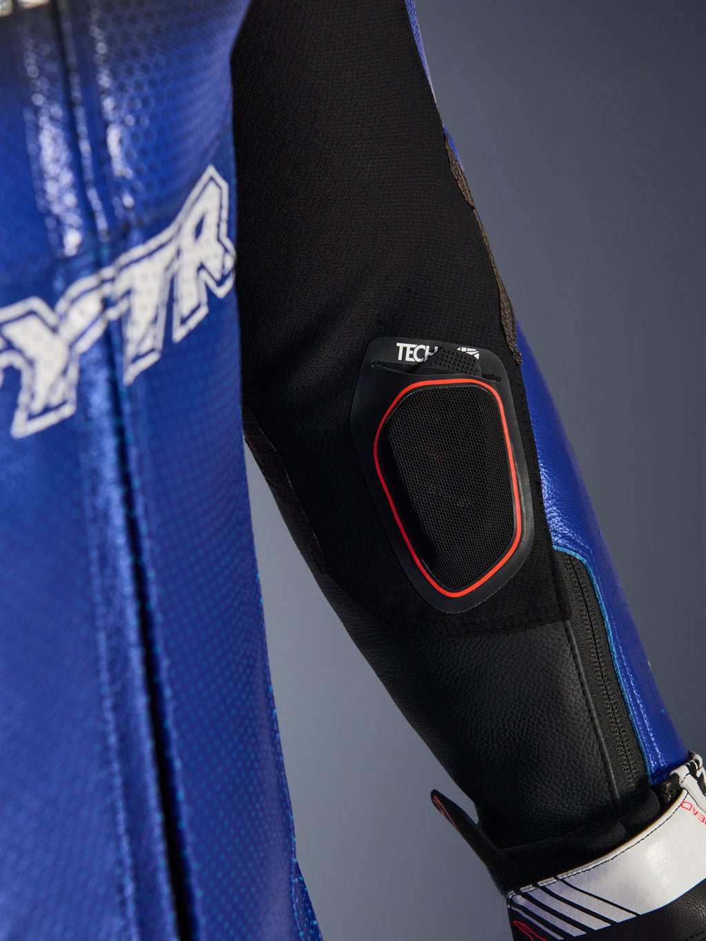 Pormenor do antebraço do fato de couro Yamaha GP-R7 One Piece, com bolso integrado para o visor Bluetooth Tech-Air 10 com contorno vermelho, couro perfurado em azul metálico e inserções de tecido elástico preto para performance em corrida.