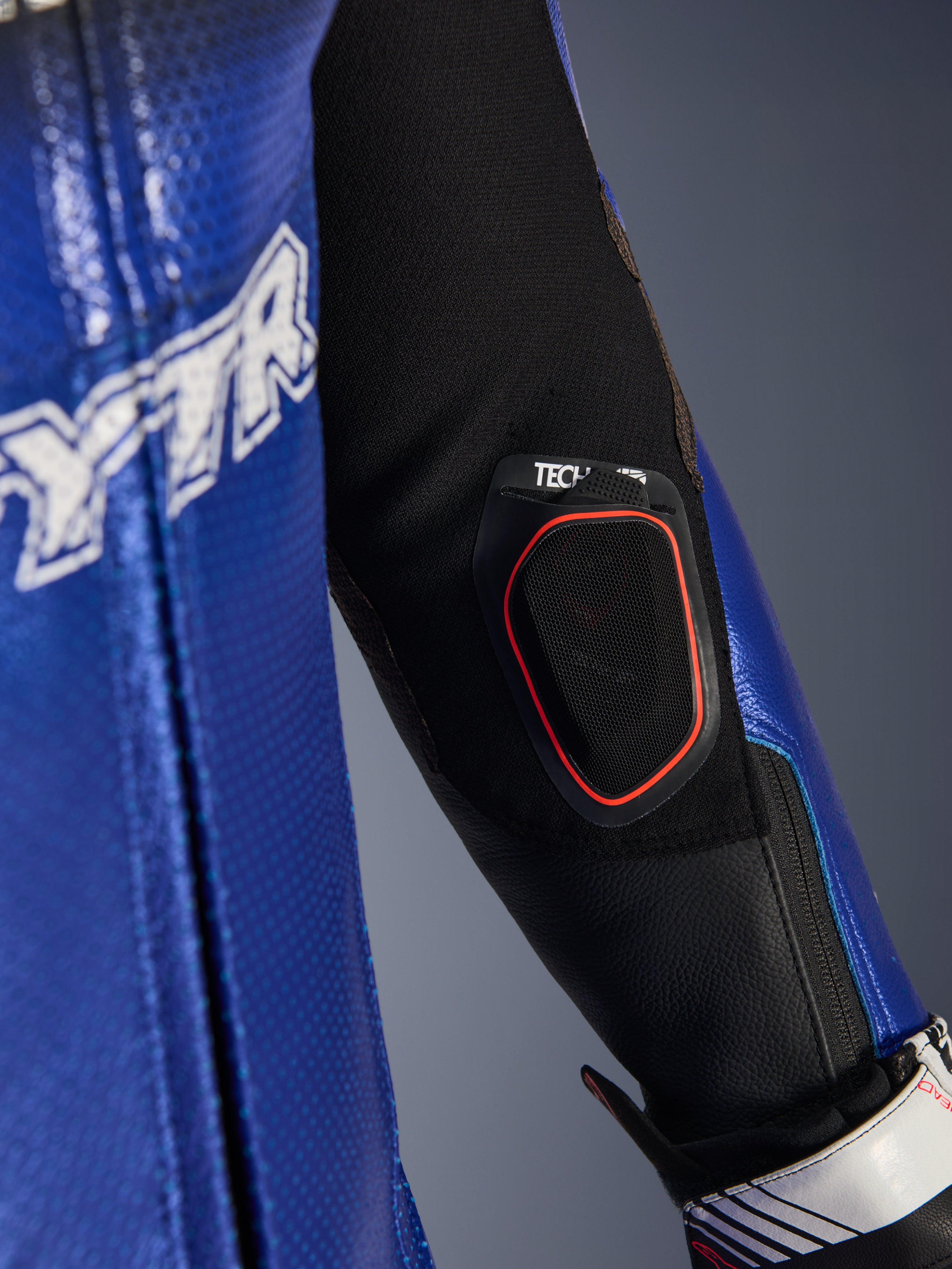 Pormenor do antebraço do fato de couro Yamaha GP-R7 One Piece, com bolso integrado para o visor Bluetooth Tech-Air 10 com contorno vermelho, couro perfurado em azul metálico e inserções de tecido elástico preto para performance em corrida.