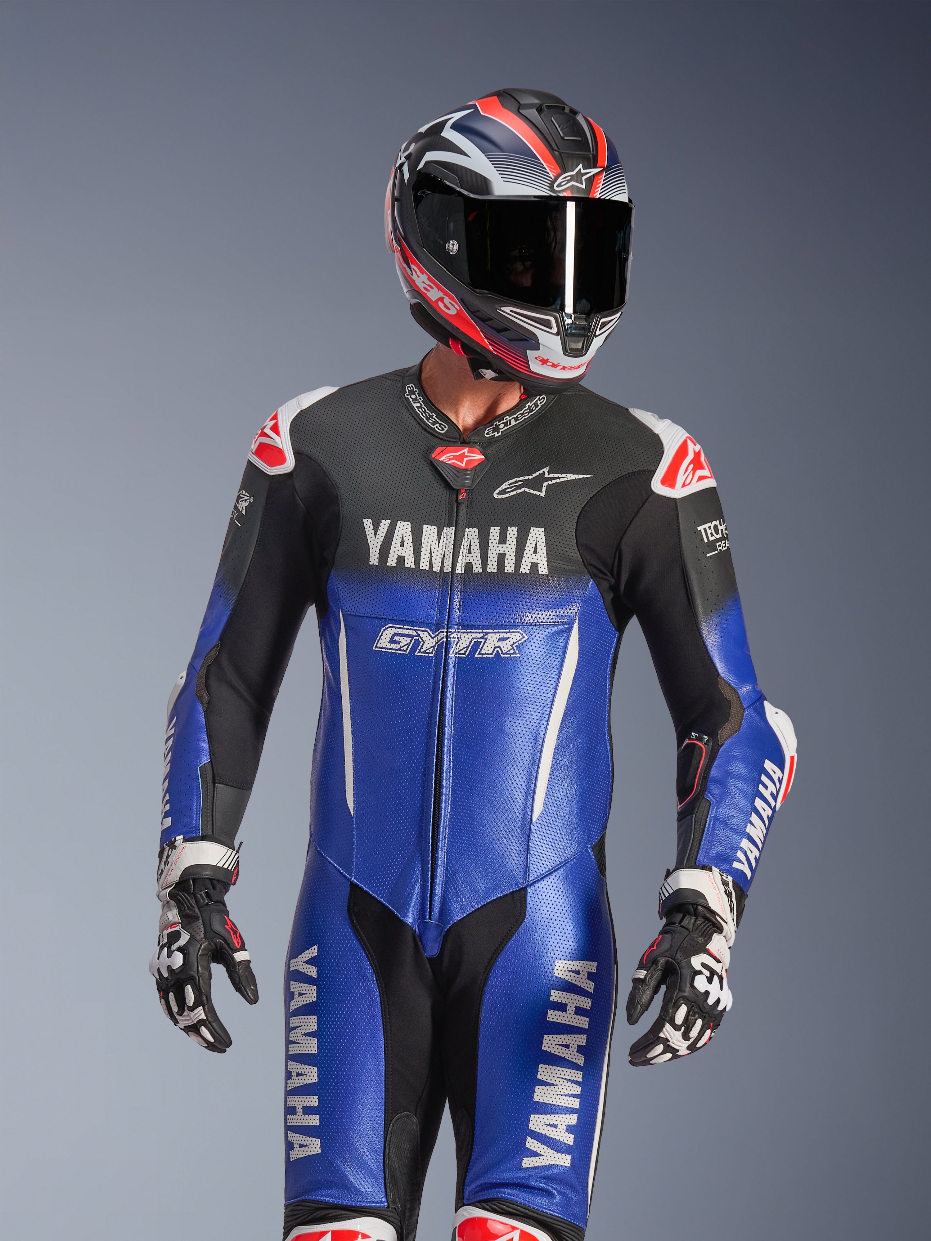 Fato de Couro Inteiro Yamaha GP-R7, azul metálico e preto com detalhes em branco e vermelho, usado com capacete e luvas de corrida a condizer, com logótipos YAMAHA e GYTR, painéis de couro perfurados e logótipos Alpinestars, vestuário profissional de motociclismo de competição.