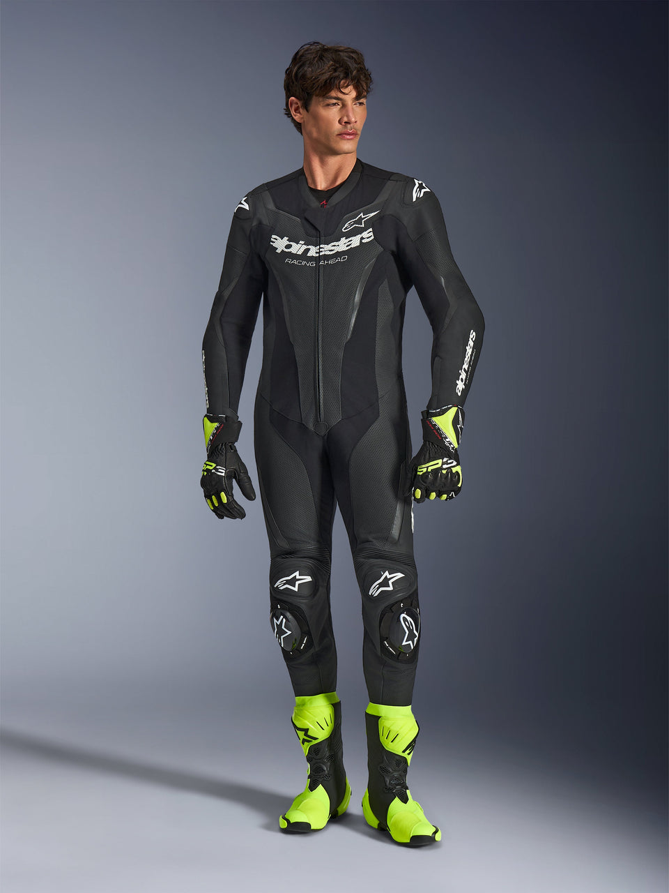 Alpinestars GP Force V2 Leather Suit, fato de corrida de motociclismo de uma peça preto, usado com botas e luvas amarelo fluorescente, com logótipos brancos e pele perfurada, design elegante e ajustado para dias de pista