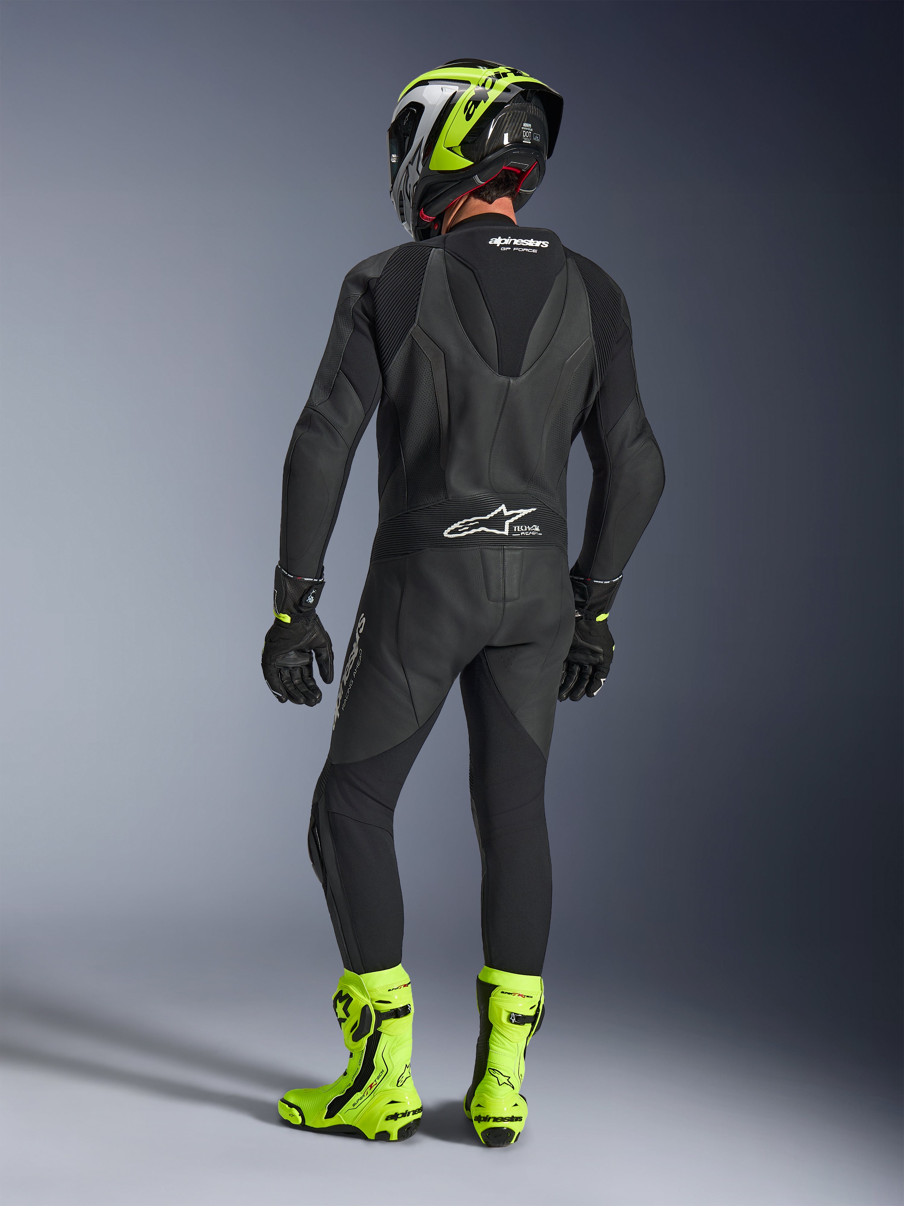 Rear view of a rider wearing the Alpinestars GP Force V2 1-Piece Leather Suit in Black, showing the aerodynamic speed hump and Tech-Air Ready branding, paired with a black and fluorescent yellow helmet and bright yellow racing boots.
