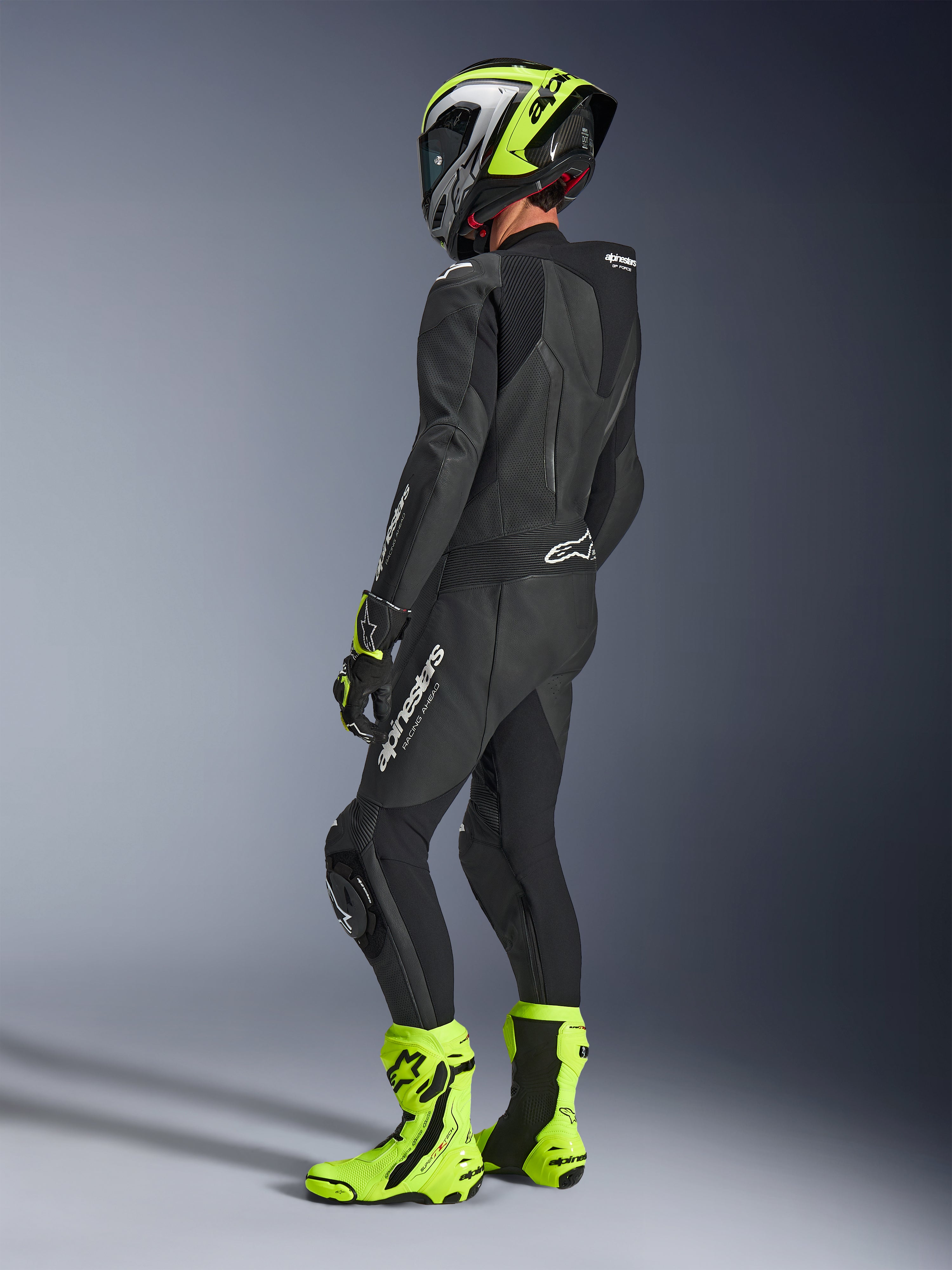 Motorcycle rider wearing Alpinestars GP Force V2 Leather Suit 1-Piece in Black, viewed from the back-side with white branding on the thigh and lower back, aerodynamic speed hump, paired with a neon yellow and black helmet, matching racing gloves and boots, professional track and sport riding gear.