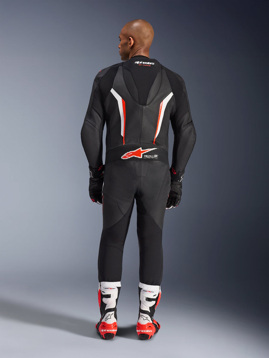 Vista traseira de um modelo a usar o Fato de Cabedal Alpinestars GP Force V2 de Uma Peça em Preto Branco Vermelho Fluo, destacando a corcova aerodinâmica e a marca Tech-Air Ready, combinado com botas de corrida profissionais brancas e vermelhas e luvas pretas.