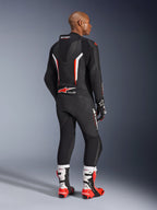Piloto a usar o Fato de Pele GP Force V2 de 1 Peça da Alpinestars, preto com detalhes em branco e vermelho fluo, visto de costas mostrando a corcova aerodinâmica e a marca Alpinestars, combinado com luvas de corrida a condizer e botas de performance brancas e vermelhas, equipamento profissional de motociclismo de competição.