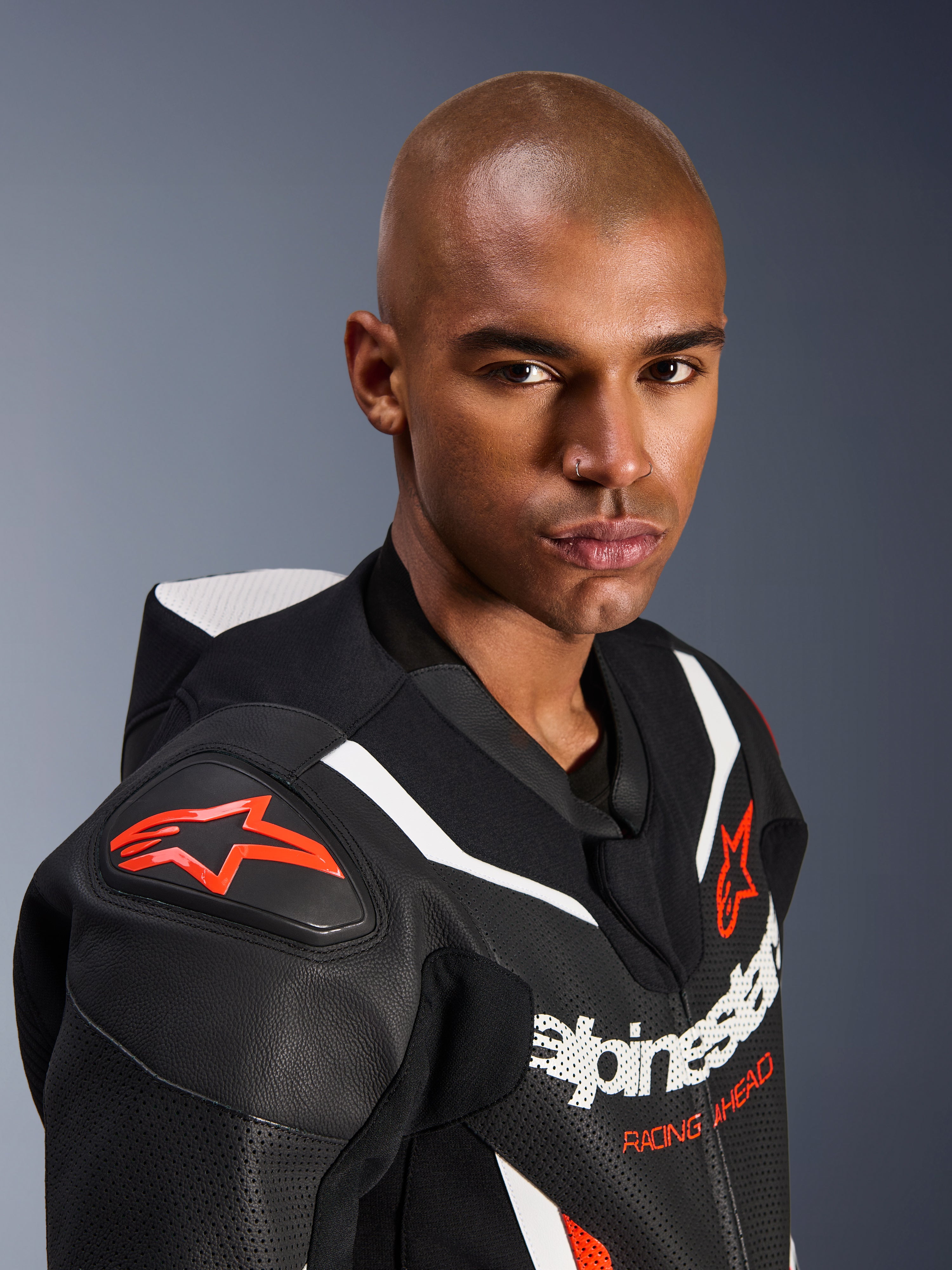 Retrato em grande plano de um modelo a usar o fato de pele Alpinestars GP Force V2 de uma peça, em preto, branco e vermelho fluo, com painéis de pele perfurada, riscas de corrida brancas e logótipos Alpinestars vermelhos no ombro e no peito.