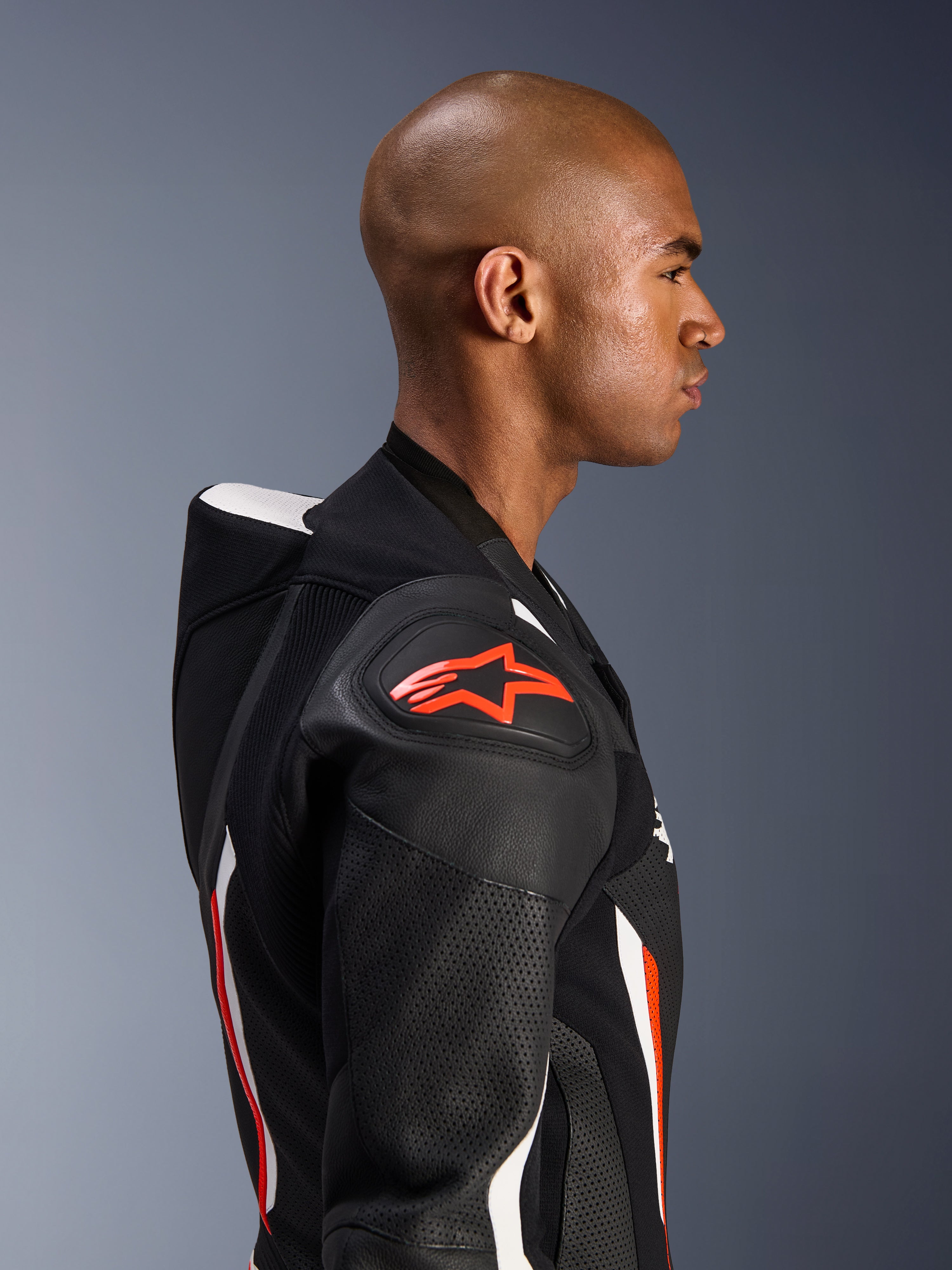 Fato de Cabedal Alpinestars GP Force V2 - 1 Peça, perfil lateral de um piloto, cores Preto Branco Vermelho Fluo, destaque para a corcova aerodinâmica, zonas de pele perfurada e proteção de ombro DFS Lite com logótipo Alpinestars vermelho
