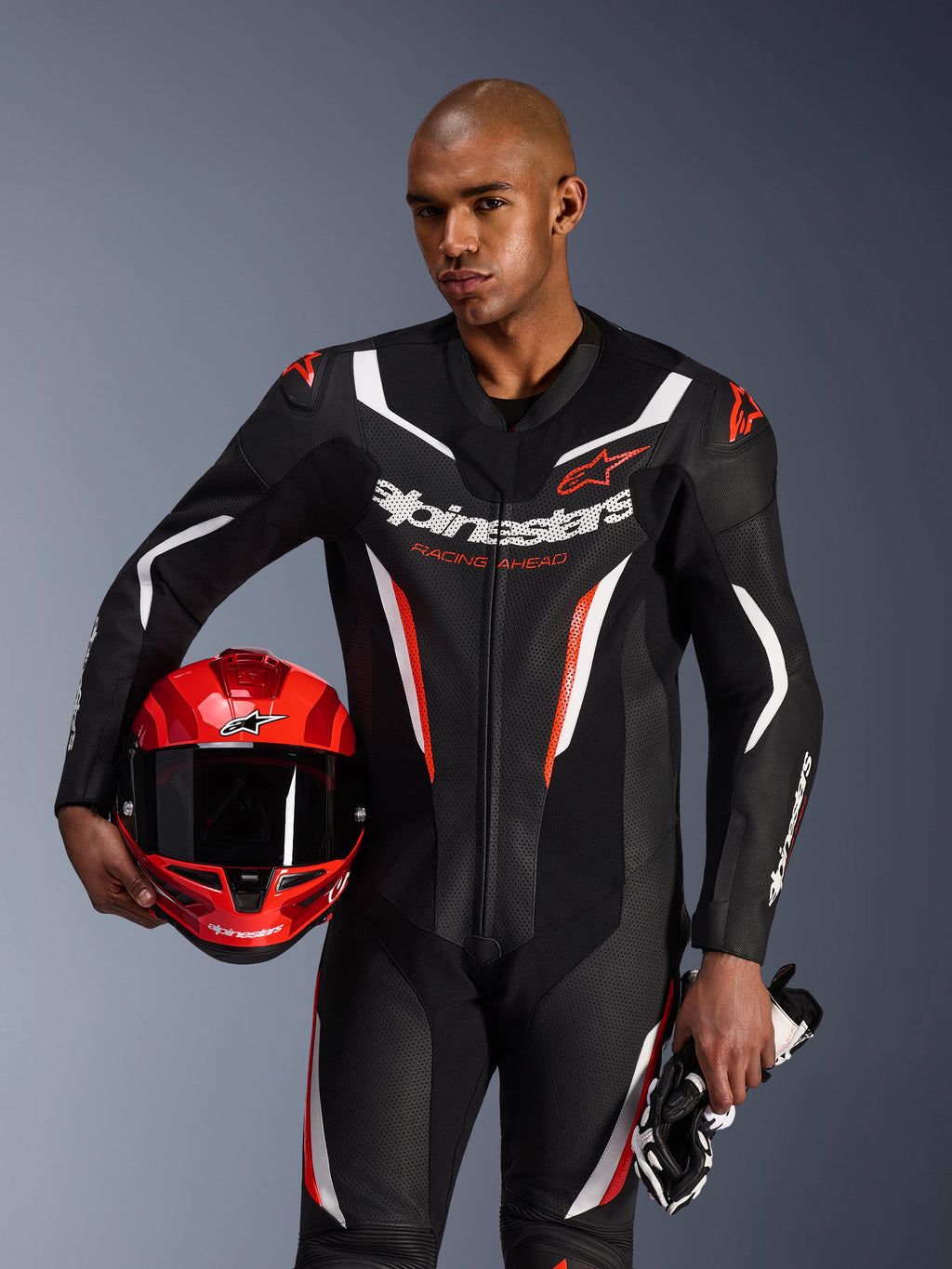 Fato de Pele Alpinestars GP Force V2, fato de motociclismo de 1 peça, preto, branco e vermelho fluo, vestido por um modelo a segurar um capacete vermelho e luvas, design de corrida elegante com pele perfurada e logótipos destacados