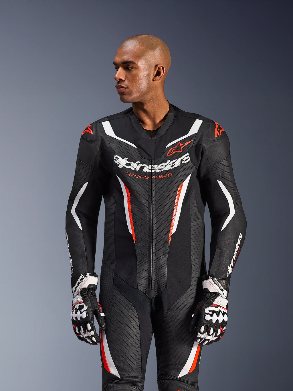 Fato de Pele Alpinestars GP Force V2 de 1 Peça, preto, branco e vermelho fluo, usado por um modelo masculino com luvas de corrida, com construção em pele perfurada, detalhes gráficos arrojados em branco e vermelho, e logótipo Alpinestars no peito.