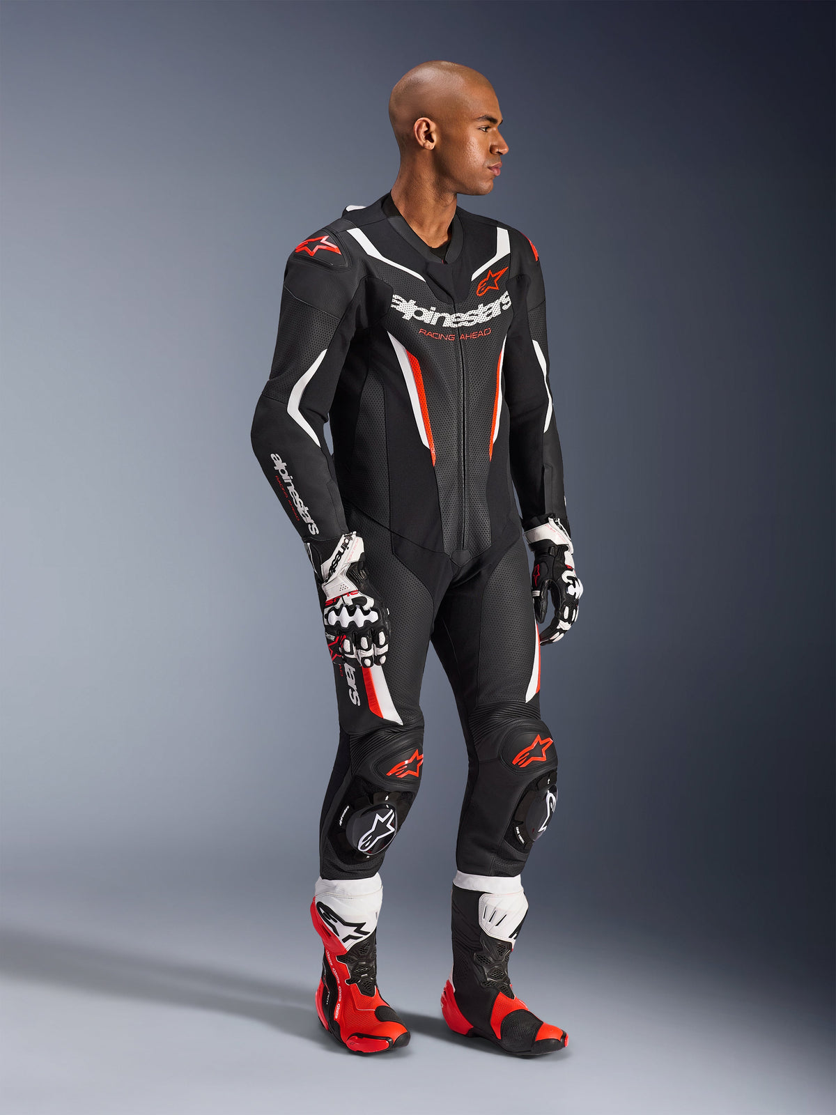 Modelo a usar o Fato de Cabedal de 1 Peça Alpinestars GP Force V2 em Preto, Branco e Vermelho Fluo, equipamento de corrida de alto desempenho com painéis de pele perfurada e proteções DFS Lite, complementado com botas e luvas de corrida em vermelho e branco a condizer, estética profissional de motociclismo.