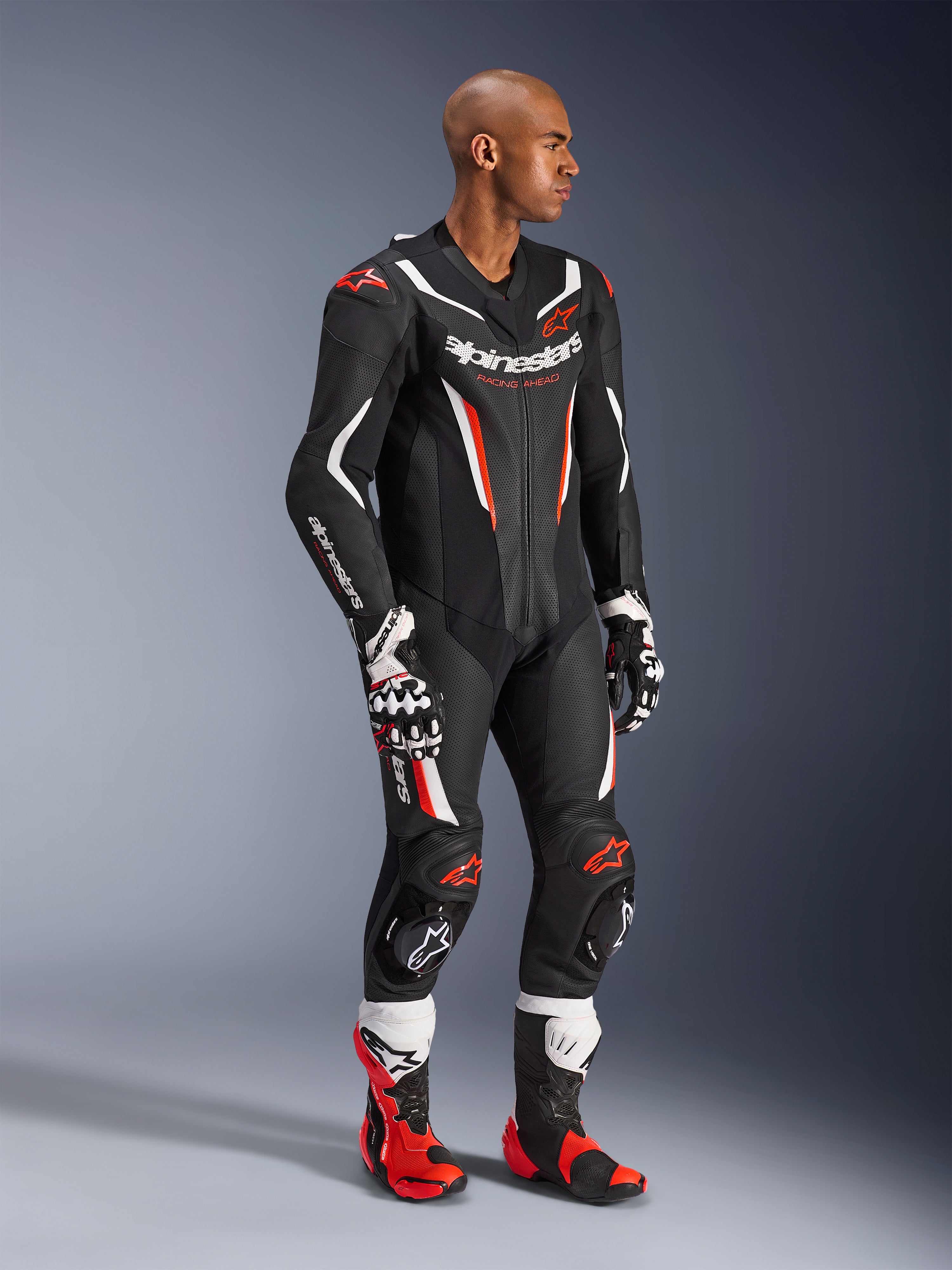 Modelo a usar o Fato de Cabedal de 1 Peça Alpinestars GP Force V2 em Preto, Branco e Vermelho Fluo, equipamento de corrida de alto desempenho com painéis de pele perfurada e proteções DFS Lite, complementado com botas e luvas de corrida em vermelho e branco a condizer, estética profissional de motociclismo.