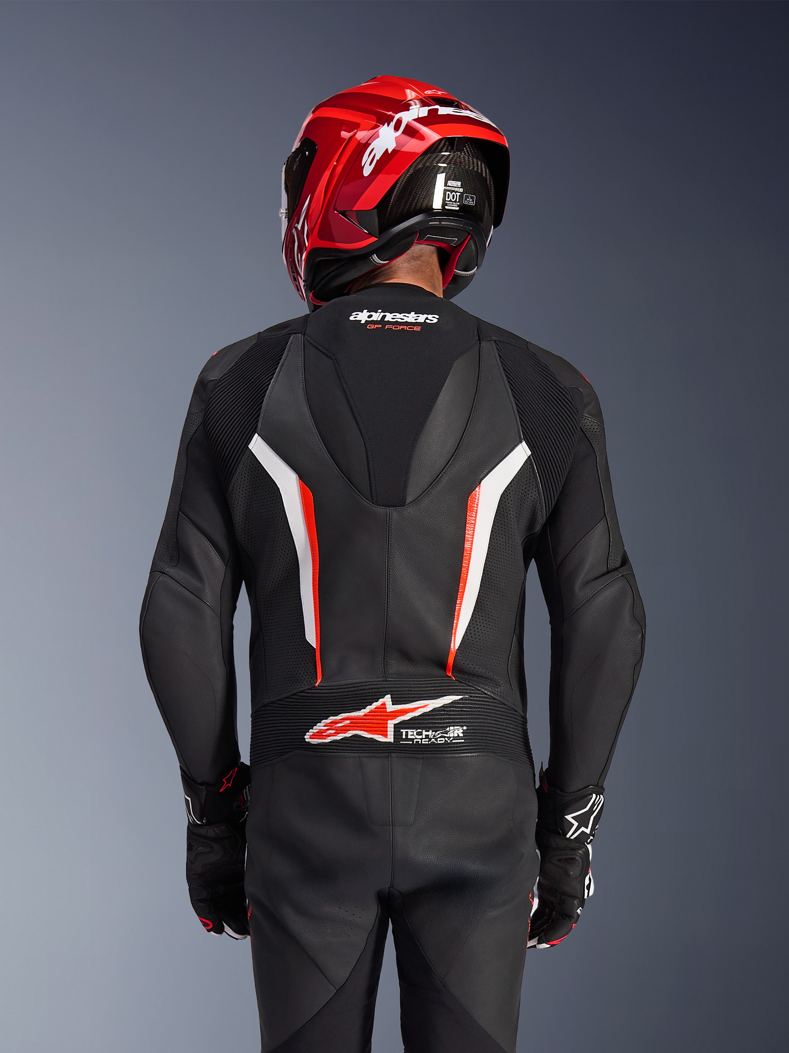 Alpinestars GP Force V2 Leather Suit 1-Piece, preto, branco e vermelho fluo, vista traseira do piloto com capacete de corrida vermelho e luvas, com painéis de pele perfurada, corcova aerodinâmica com marca Alpinestars e logótipo Tech-Air Ready na zona lombar, equipamento profissional de motociclismo