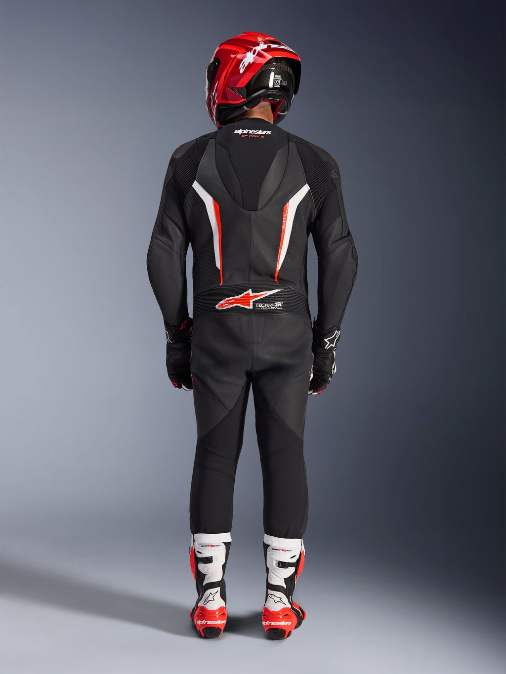 Piloto visto de costas a usar o Fato de Cabedal de 1 Peça Alpinestars GP Force V2 em Preto Branco Vermelho Fluo, com corcova aerodinâmica e marcação Tech-Air Ready, coordenado com um capacete de corrida vermelho e botas brancas e vermelhas, design de equipamento de pista profissional.