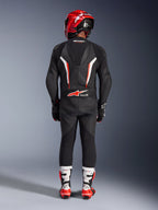 Piloto visto de costas a usar o Fato de Cabedal de 1 Peça Alpinestars GP Force V2 em Preto Branco Vermelho Fluo, com corcova aerodinâmica e marcação Tech-Air Ready, coordenado com um capacete de corrida vermelho e botas brancas e vermelhas, design de equipamento de pista profissional.
