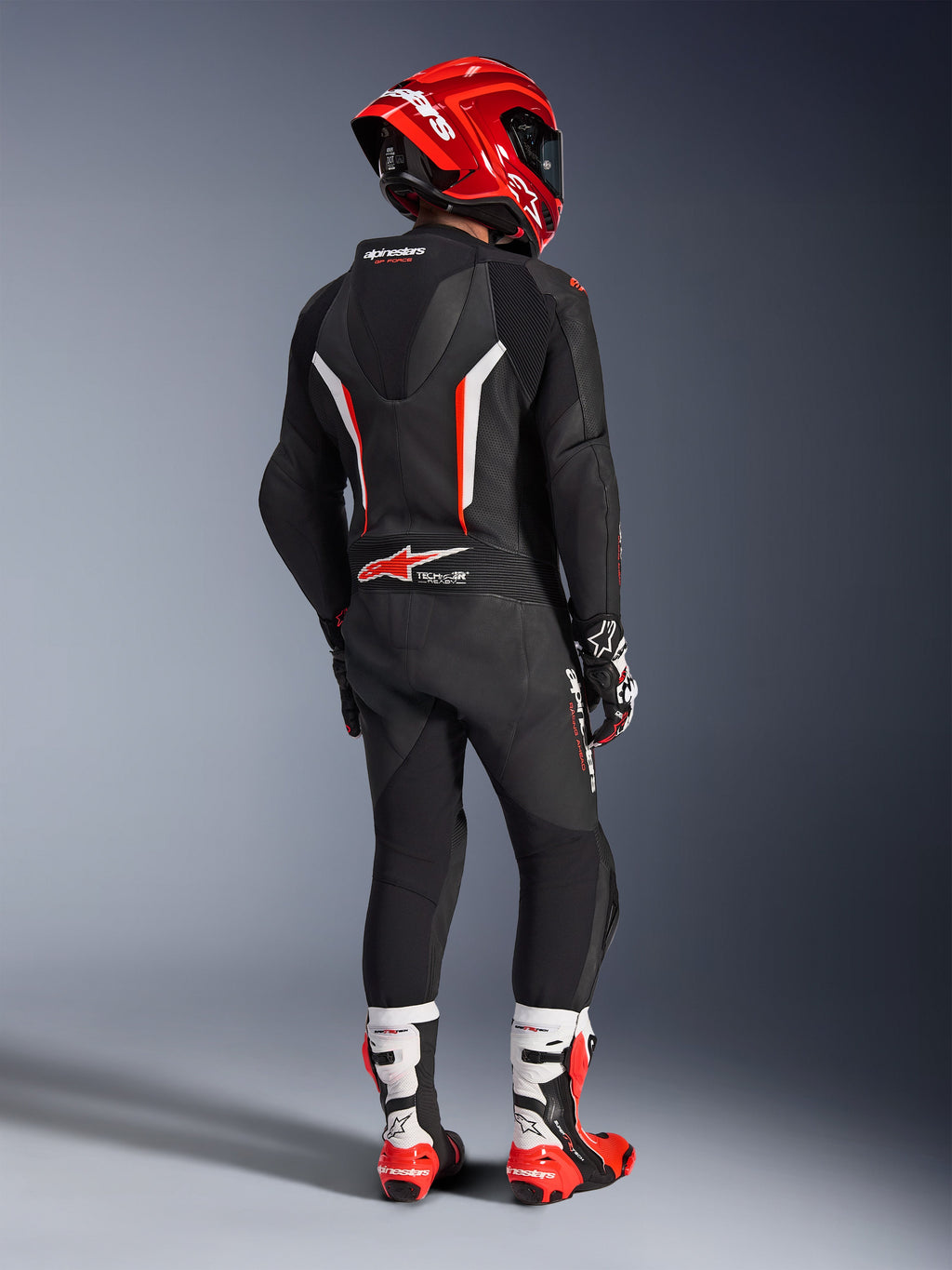 Piloto visto de costas com o Fato de Pele de uma peça Alpinestars GP Force V2 em preto, branco e vermelho fluo, com corcova aerodinâmica e painéis de pele perfurada, acompanhado por um capacete Alpinestars vermelho, luvas de corrida e botas de motociclismo vermelhas e brancas, equipamento profissional de pista.