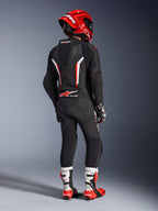 Piloto visto de costas com o Fato de Pele de uma peça Alpinestars GP Force V2 em preto, branco e vermelho fluo, com corcova aerodinâmica e painéis de pele perfurada, acompanhado por um capacete Alpinestars vermelho, luvas de corrida e botas de motociclismo vermelhas e brancas, equipamento profissional de pista.