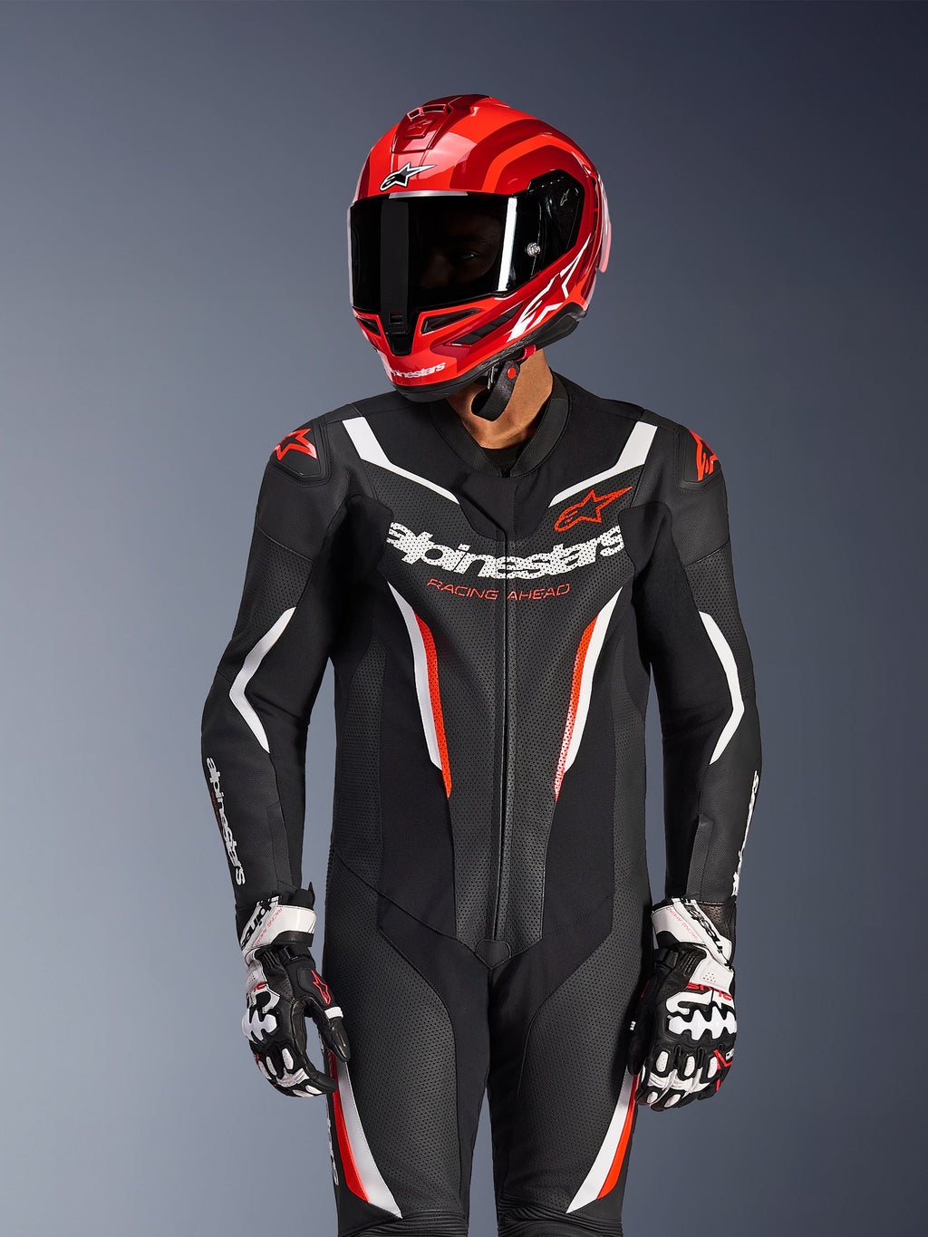 Motociclista a usar o fato de pele Alpinestars GP Force V2 de uma peça em preto, branco e vermelho fluo, capacete de corrida e luvas em vermelho coordenado, apresenta painéis de pele perfurada e logótipos em destaque, equipamento de corrida de alta performance