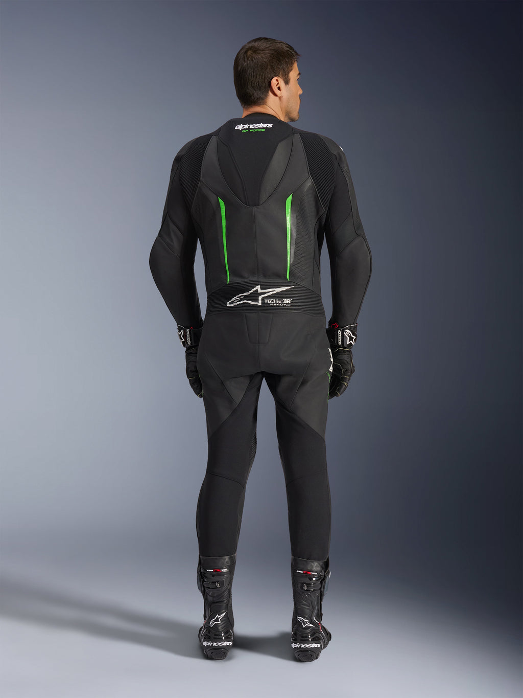 Vista traseira do Fato de Pele Alpinestars GP Force V2 de 1 Peça em preto e verde, com corcova aerodinâmica, painéis perfurados e logótipo Tech-Air Ready na zona lombar, usado com luvas e botas de corrida pretas.