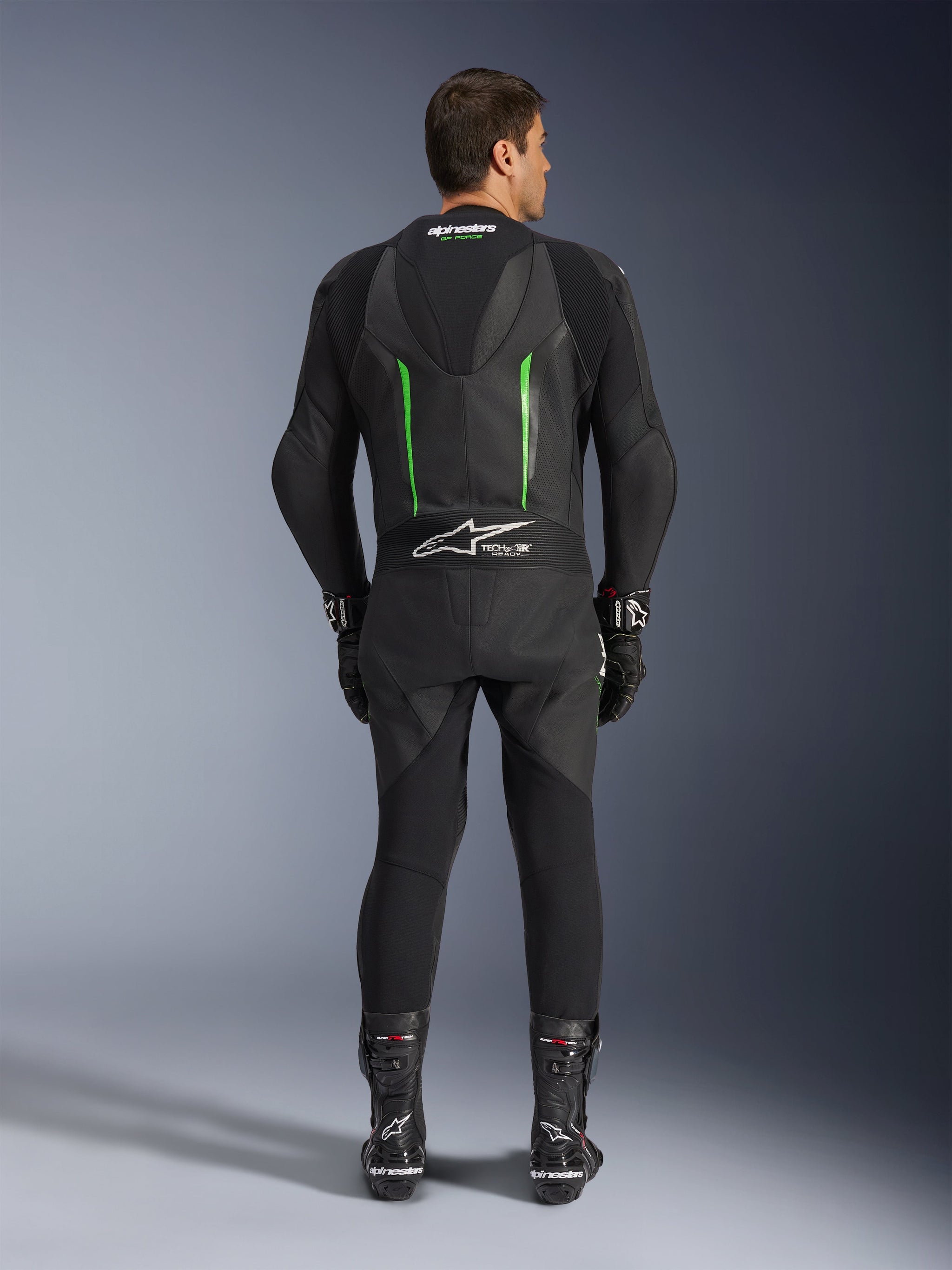 Vista traseira do Fato de Pele Alpinestars GP Force V2 de 1 Peça em preto e verde, com corcova aerodinâmica, painéis perfurados e logótipo Tech-Air Ready na zona lombar, usado com luvas e botas de corrida pretas.