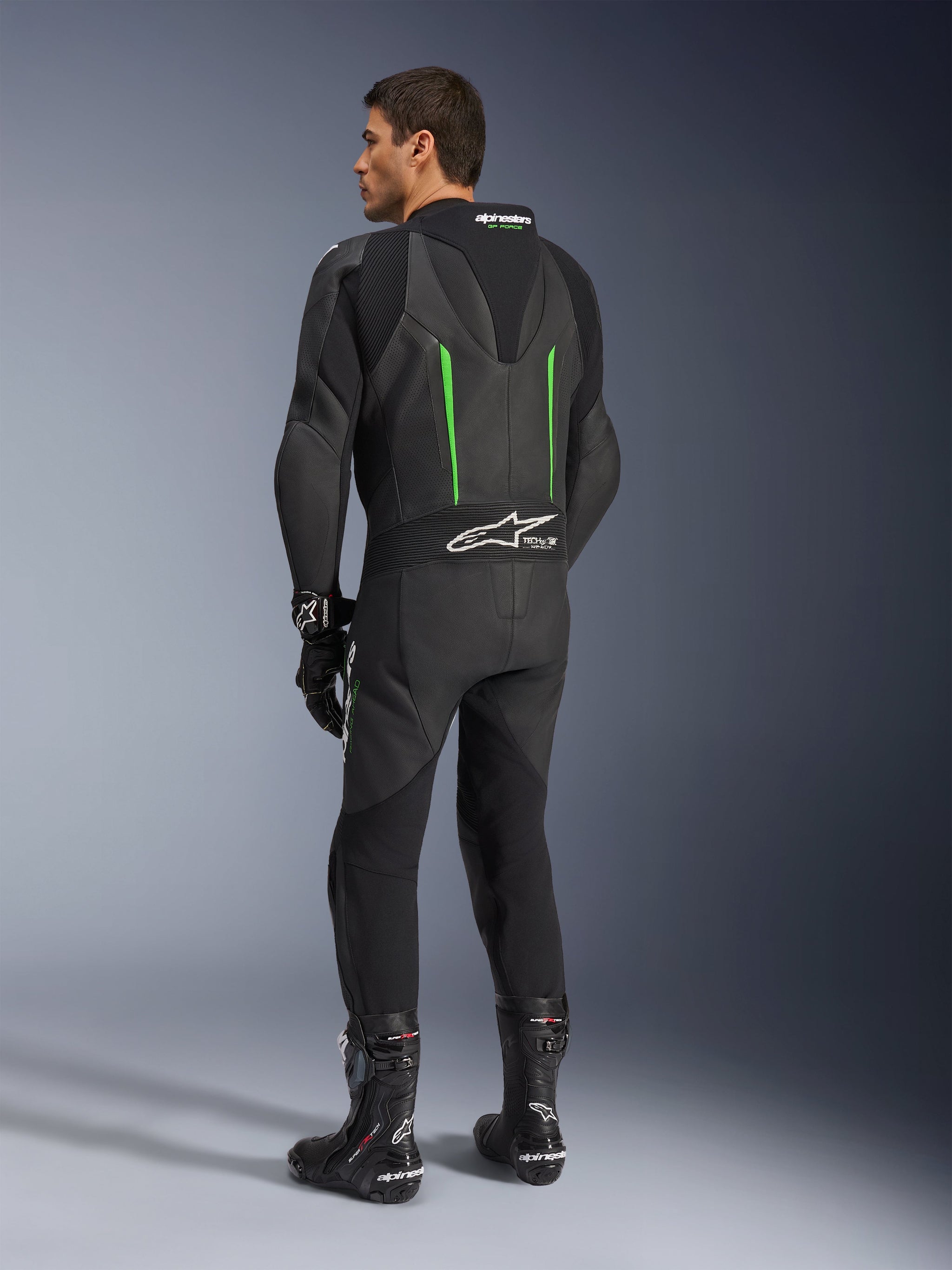 Alpinestars GP Force V2 Leather Suit, fato de motociclismo de 1 peça, preto com pormenores verdes, vista traseira mostrando a corcova aerodinâmica e o logótipo branco no painel de pele sanfonada, combinado com botas e luvas de corrida pretas.