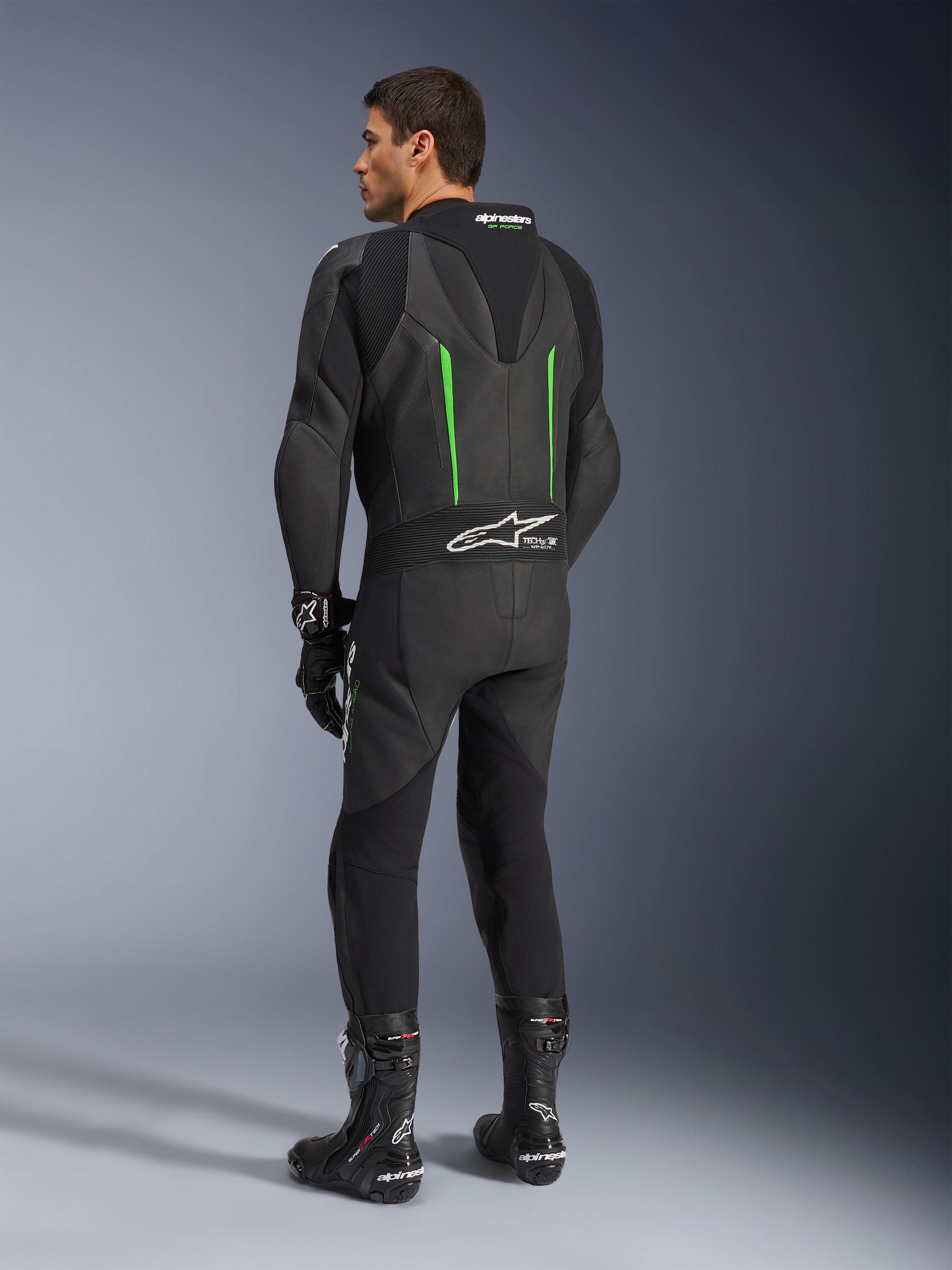 Alpinestars GP Force V2 Leather Suit, fato de motociclismo de 1 peça, preto com pormenores verdes, vista traseira mostrando a corcova aerodinâmica e o logótipo branco no painel de pele sanfonada, combinado com botas e luvas de corrida pretas.