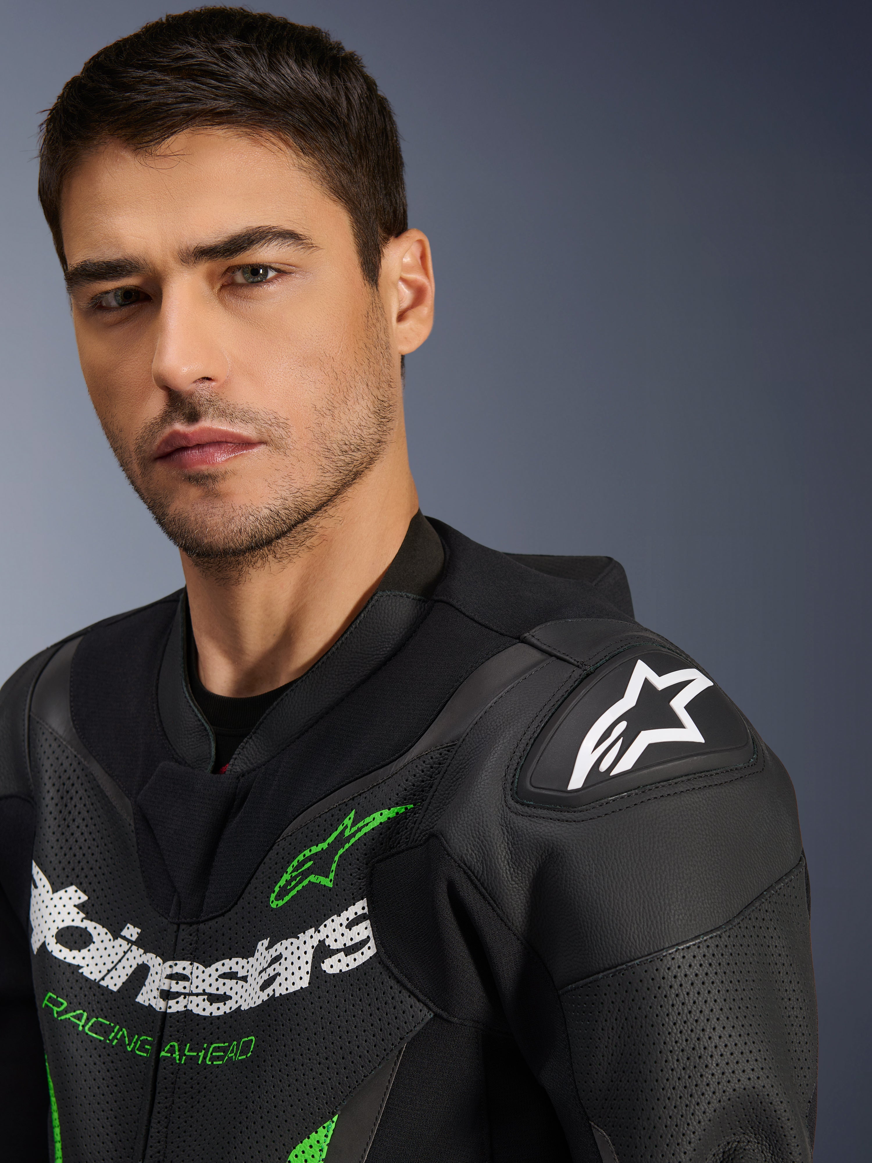 Fato de Pele Alpinestars GP Force V2 - 1 Peça, preto e verde, grande plano de um modelo masculino a usar o fato de corrida para motociclista, com logótipos em branco e verde, painéis de pele perfurada e proteção de ombro DFS Lite com logótipo de estrela branco.