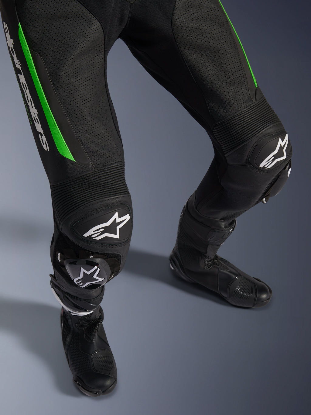Fato de Pele Alpinestars GP Force V2 - 1 Peça, grande plano das pernas em preto e verde, com construção em pele perfurada, painéis de pele sanfonada nos joelhos, logótipo Alpinestars em branco e deslizadores de joelho integrados, usado com botas de corrida pretas