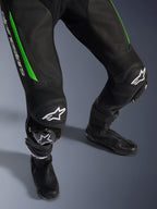 Fato de Pele Alpinestars GP Force V2 - 1 Peça, grande plano das pernas em preto e verde, com construção em pele perfurada, painéis de pele sanfonada nos joelhos, logótipo Alpinestars em branco e deslizadores de joelho integrados, usado com botas de corrida pretas
