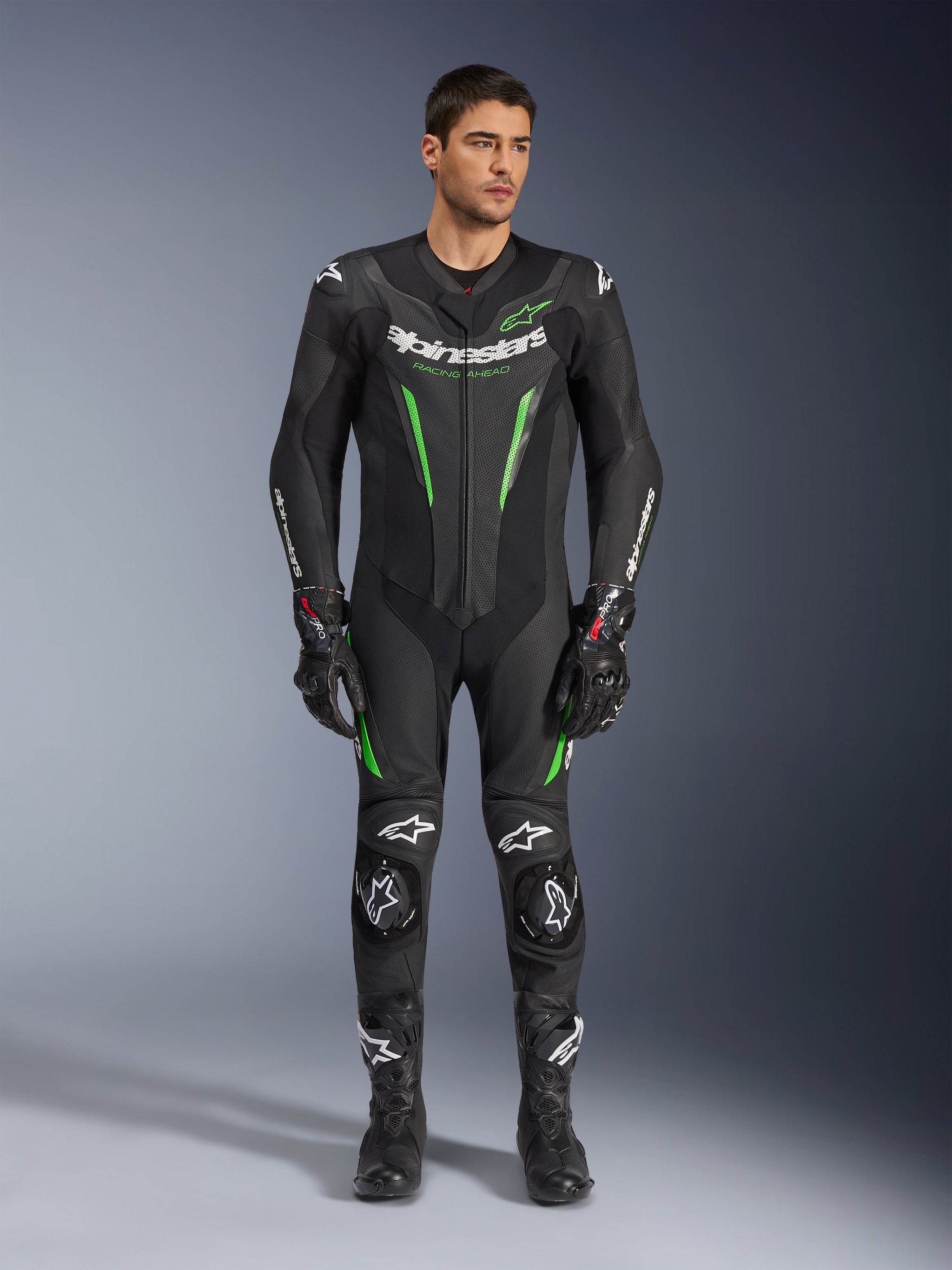 Alpinestars GP Force V2 Leather Suit - 1Piece, fato de motociclismo de uma peça, preto e verde, vista frontal de um modelo a usar o fato com logótipo branco no peito e detalhes em verde néon, com pele perfurada e deslizadores externos nos ombros, combinado com luvas e botas de corrida pretas