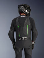 Vista traseira de um piloto com o Fato de Pele Alpinestars GP Force V2 em Preto e Verde, apresentando uma corcova aerodinâmica, o logótipo Tech-Air Ready no painel sanfonado na parte inferior das costas e construção em pele perfurada, acompanhado por um capacete de corrida em fibra de carbono preto.