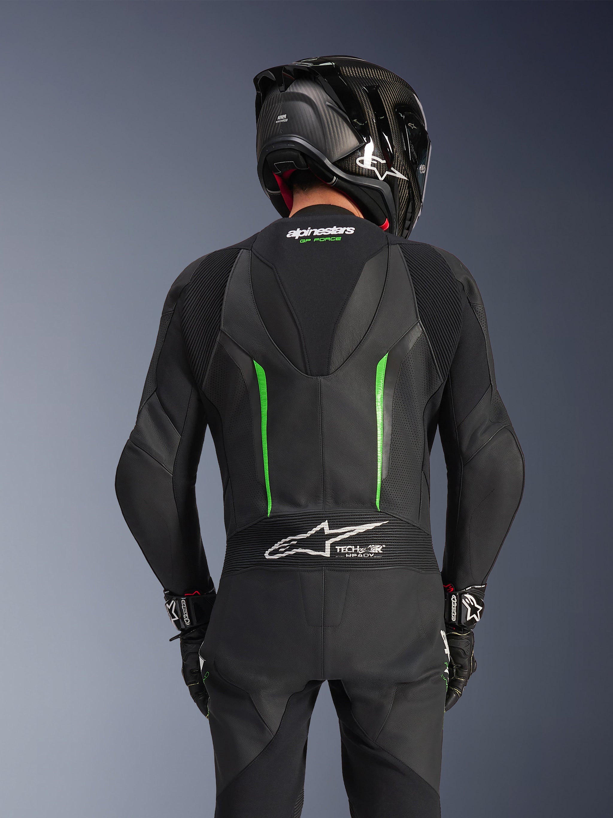 Vista traseira de um piloto com o Fato de Pele Alpinestars GP Force V2 em Preto e Verde, apresentando uma corcova aerodinâmica, o logótipo Tech-Air Ready no painel sanfonado na parte inferior das costas e construção em pele perfurada, acompanhado por um capacete de corrida em fibra de carbono preto.